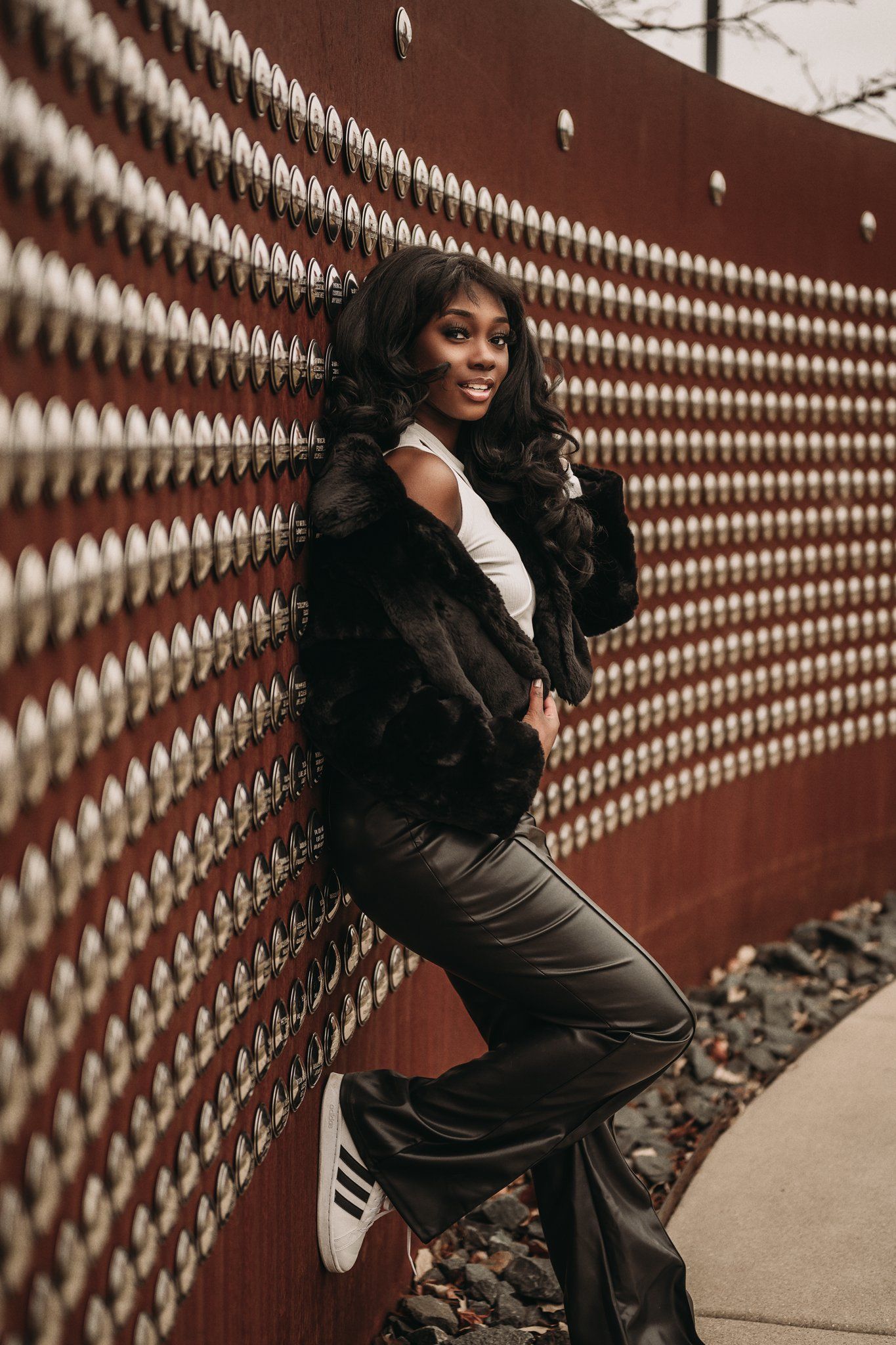 Model poses in black fur coat and leather pants against textured red wall with geometric pattern.