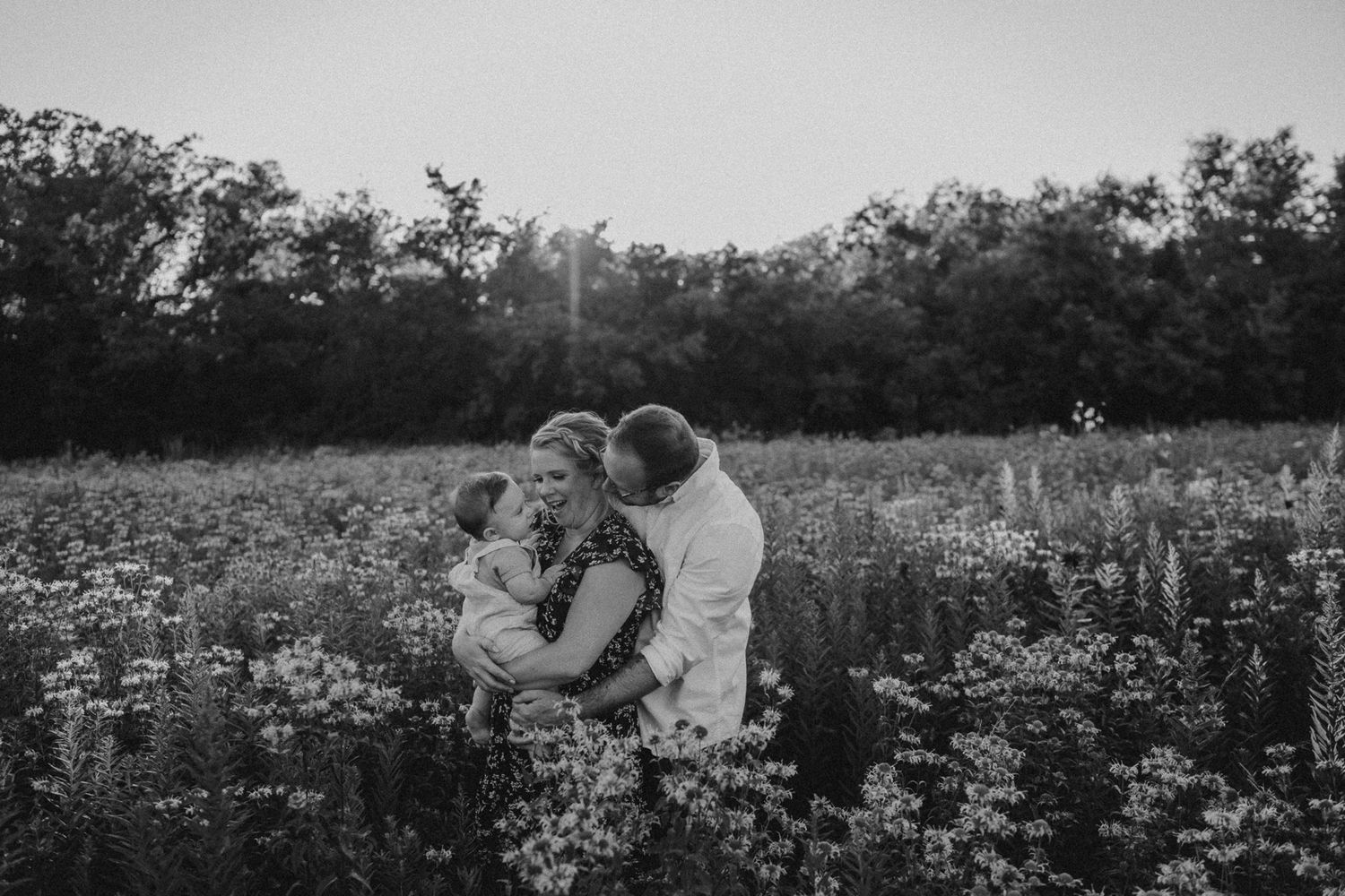 A couple shares an intimate moment in a wildflower meadow at sunset, surrounded by blooming flowers and trees.