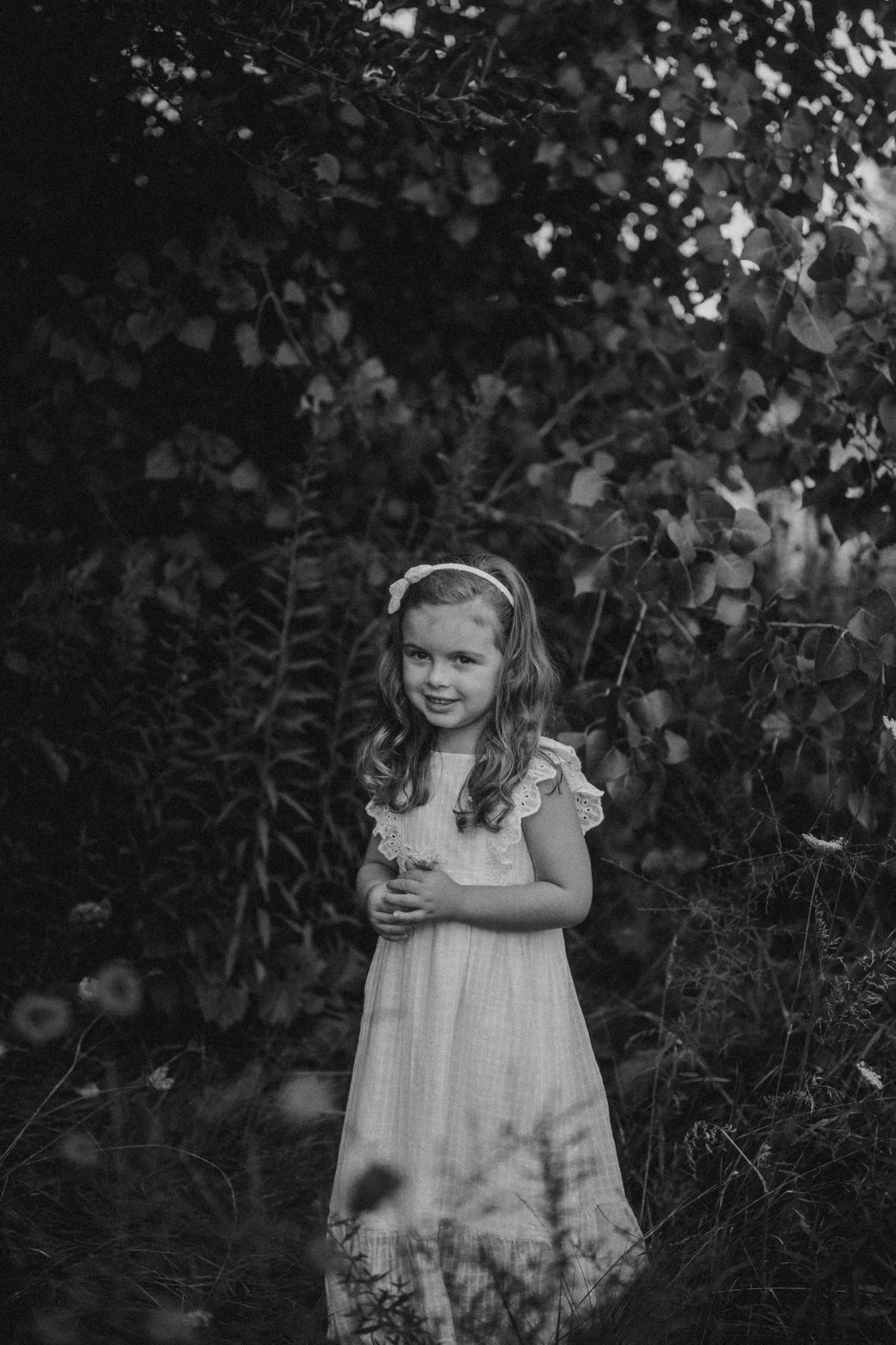 A child in a light dress poses in black and white portrait style against natural greenery.