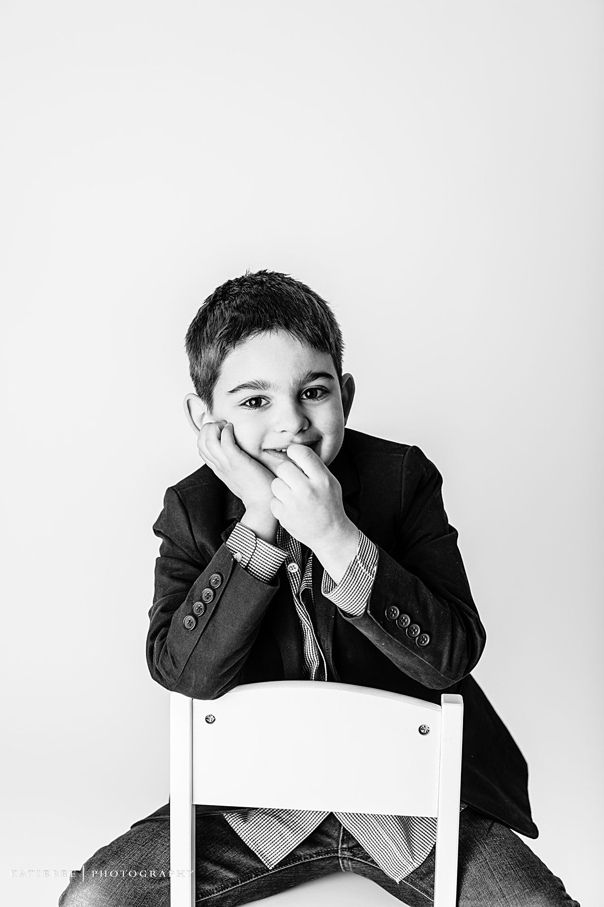 Monochrome image of child in suit jacket resting head on hand while seated in white chair.