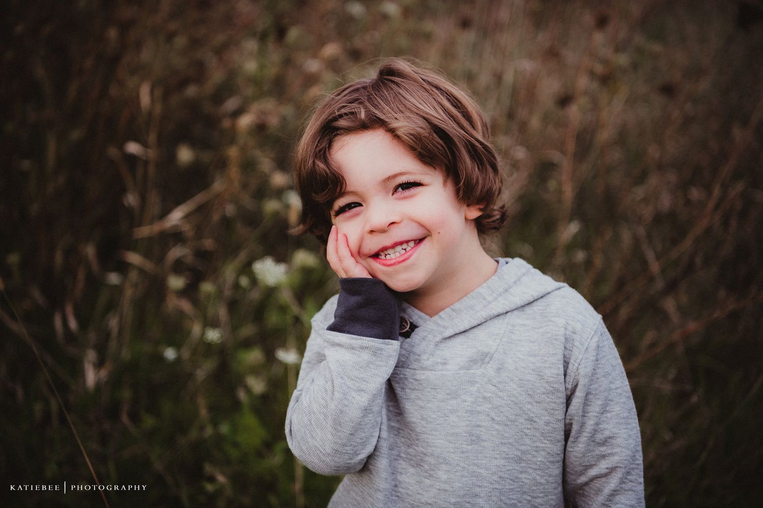 Child in grey sweatshirt smiling outdoors against natural background.