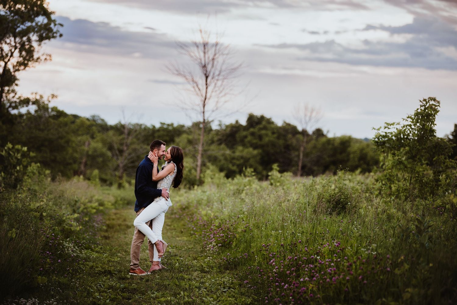 Couple embracing in a dreamy meadow setting with dramatic cloudy sky overhead.