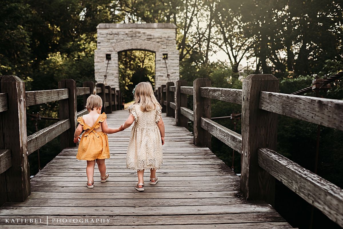Two children walking hand in hand on a wooden bridge towards a stone archway.