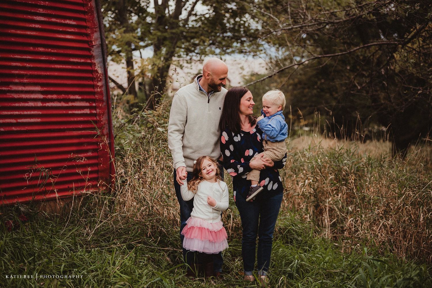 A family shares a candid moment outside a rustic red barn in autumn.