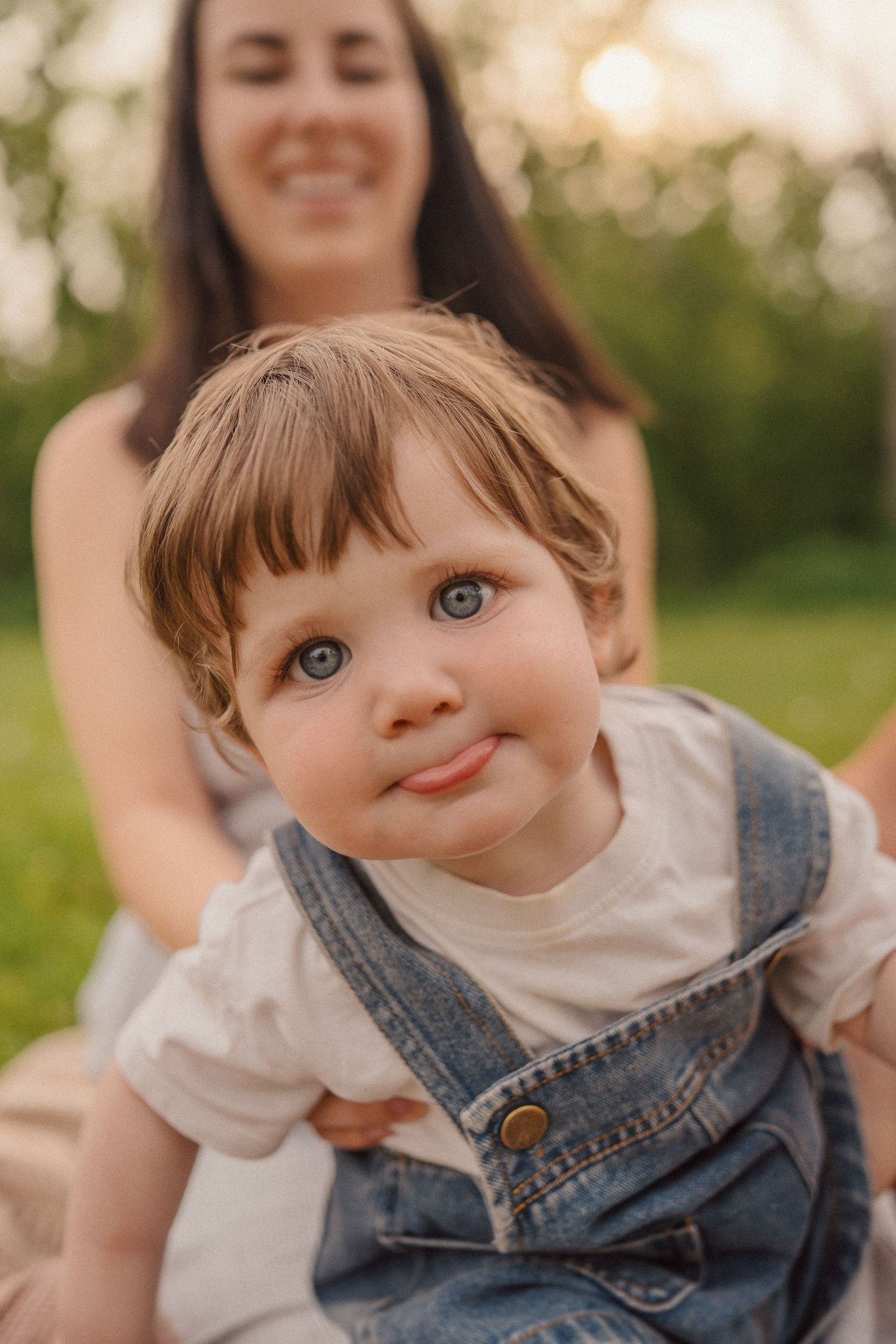 Sweet toddler wearing denim overalls smiles at camera while sitting outside during golden hour.