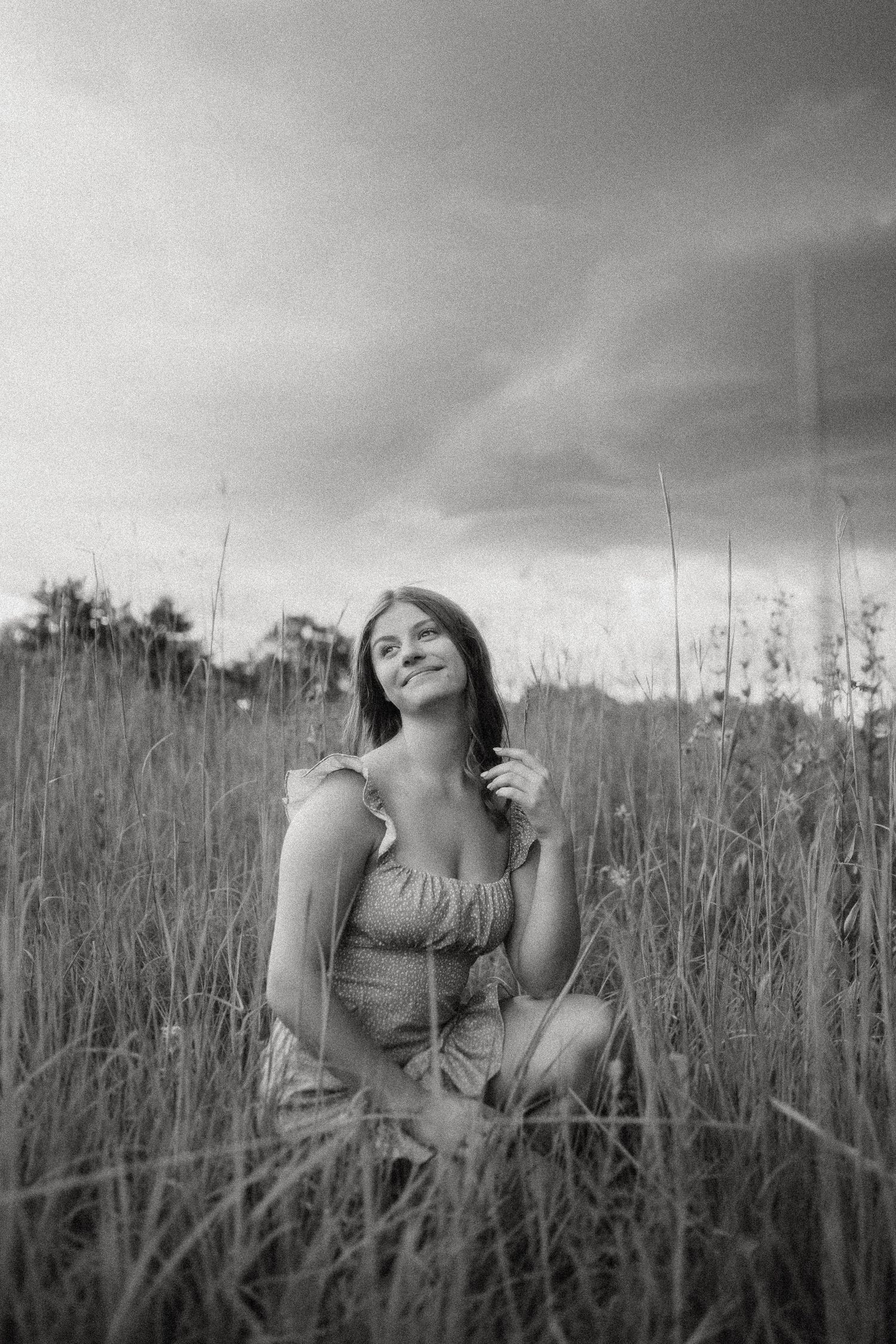 Black and white portrait of a person sitting in tall grass field under dramatic cloudy sky.