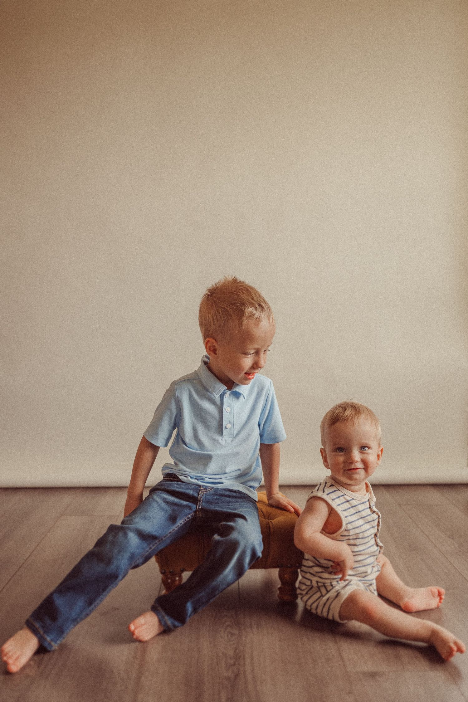 Two children sit together on hardwood flooring against a neutral wall background.