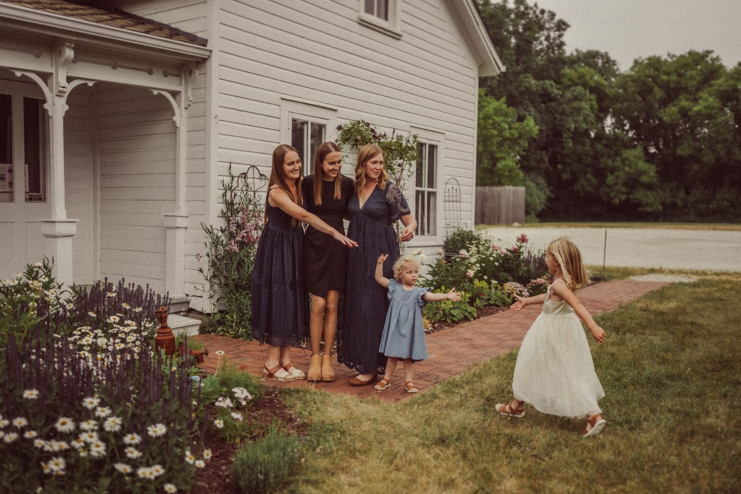 People gather on a white farmhouse porch while children play in a flower garden.