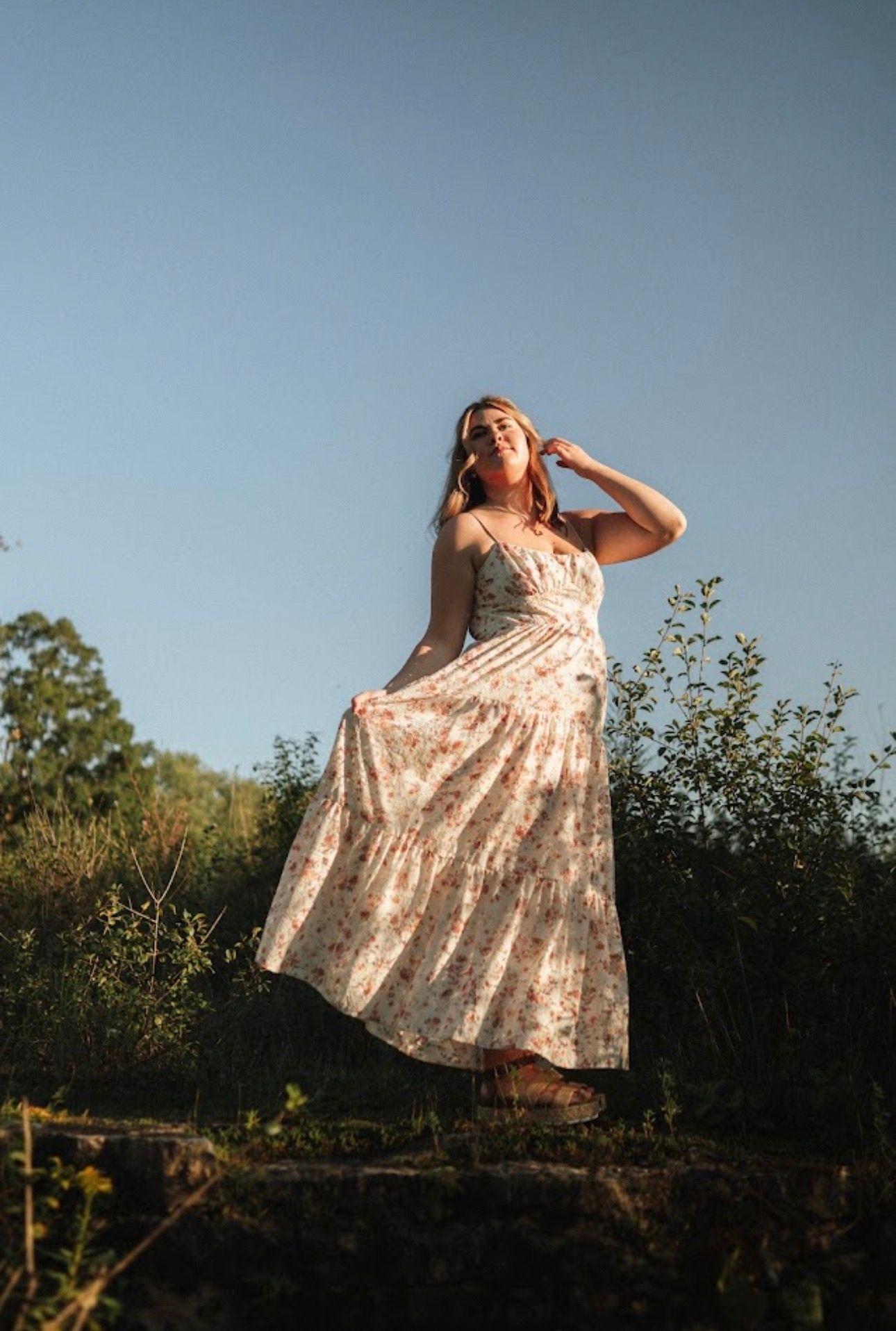 Ethereal portrait of someone in a floral dress posed against a bright sky during golden hour.
