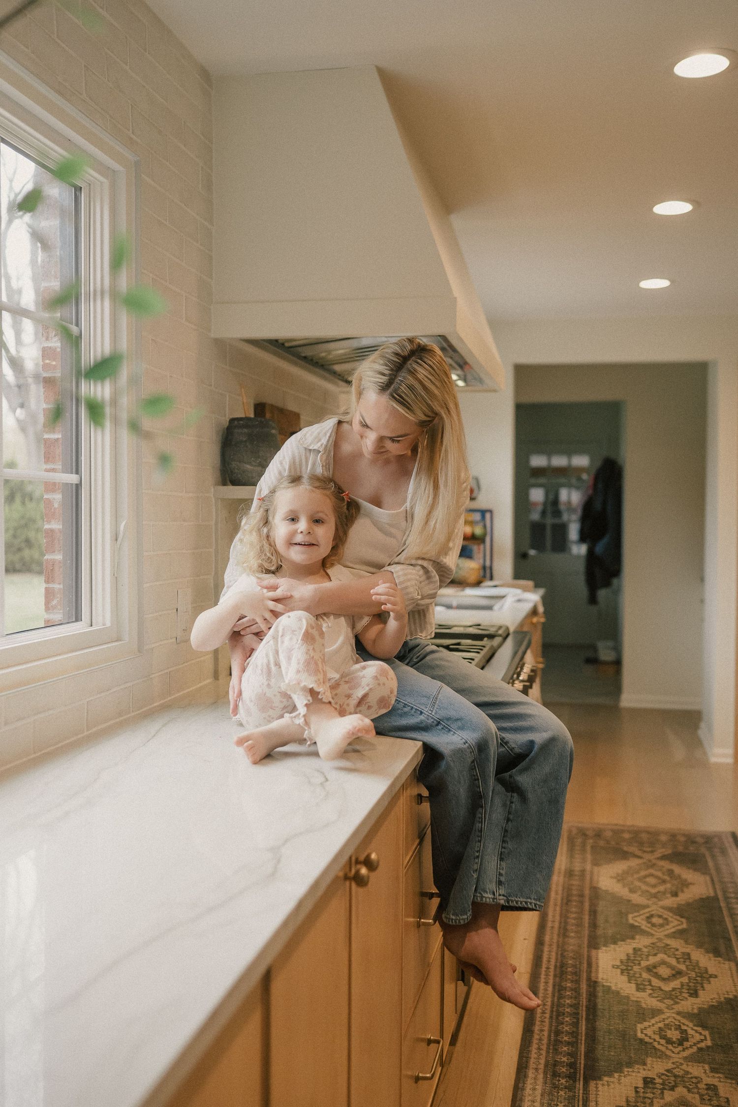 A mother sits on a kitchen counter hugging her young daughter in a bright, modern kitchen with wood cabinets.