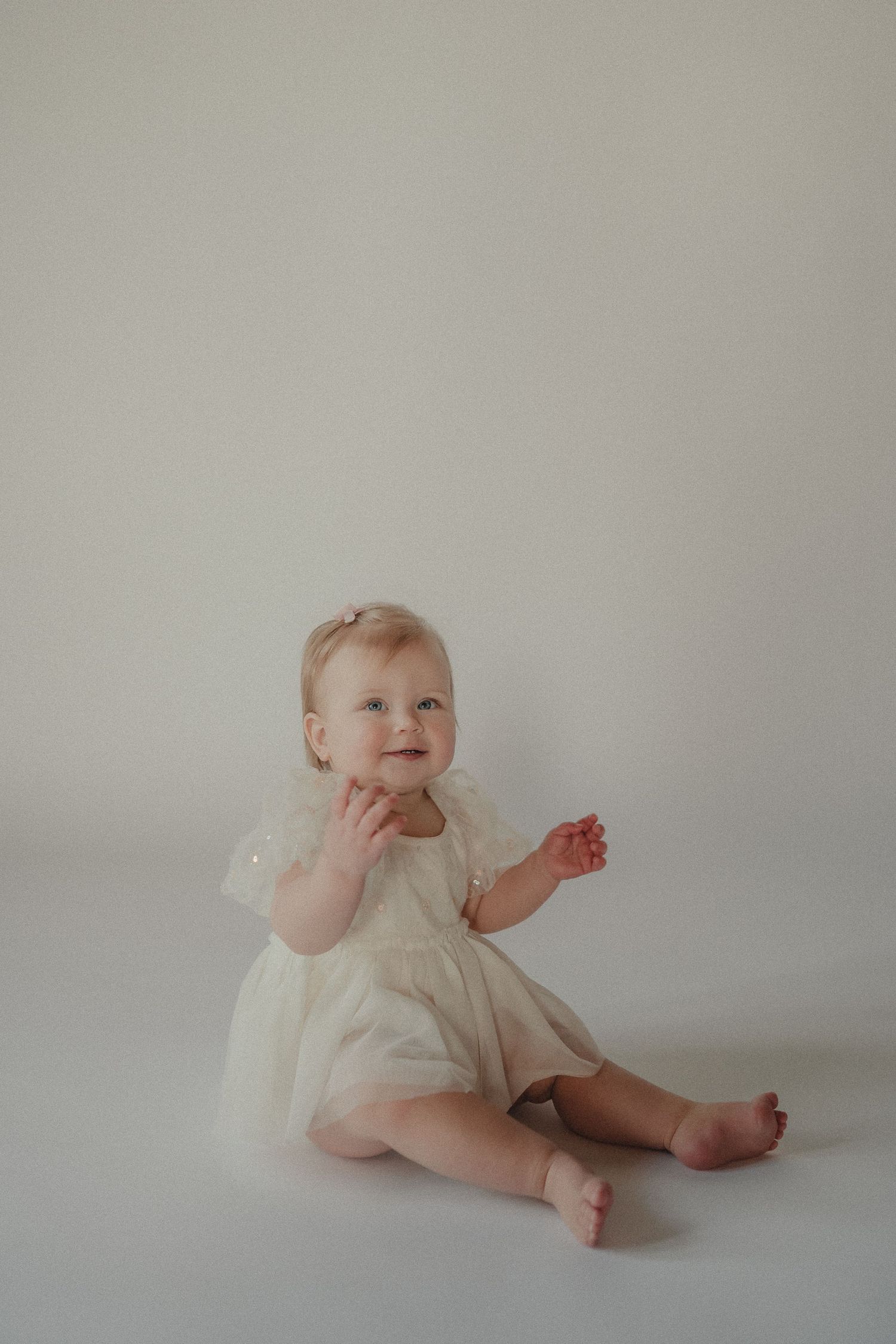 Happy baby girl in cream tulle dress clapping hands while sitting on white studio floor.