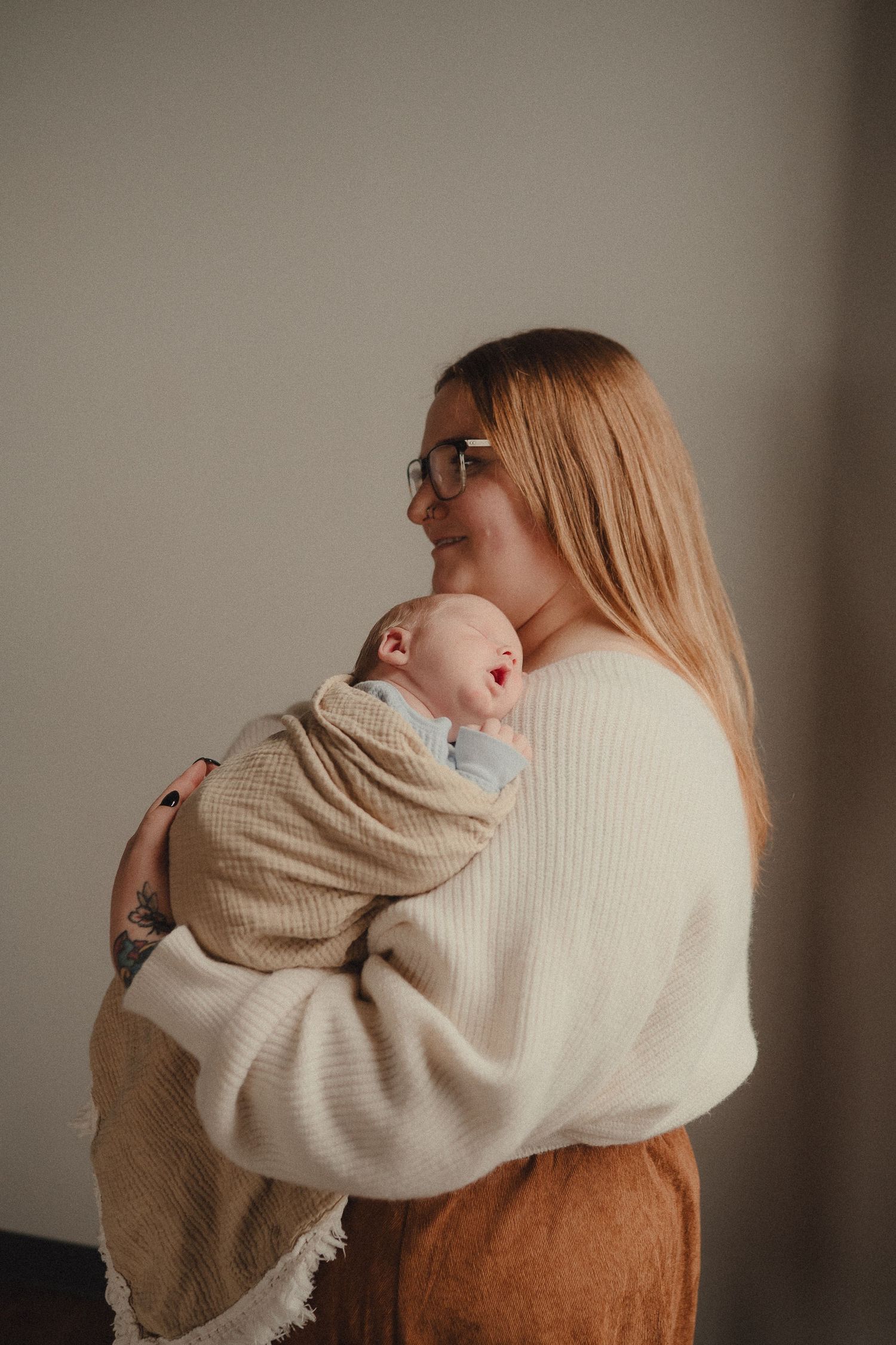 Smiling mother in cream sweater holds swaddled newborn baby against neutral wall in soft light.