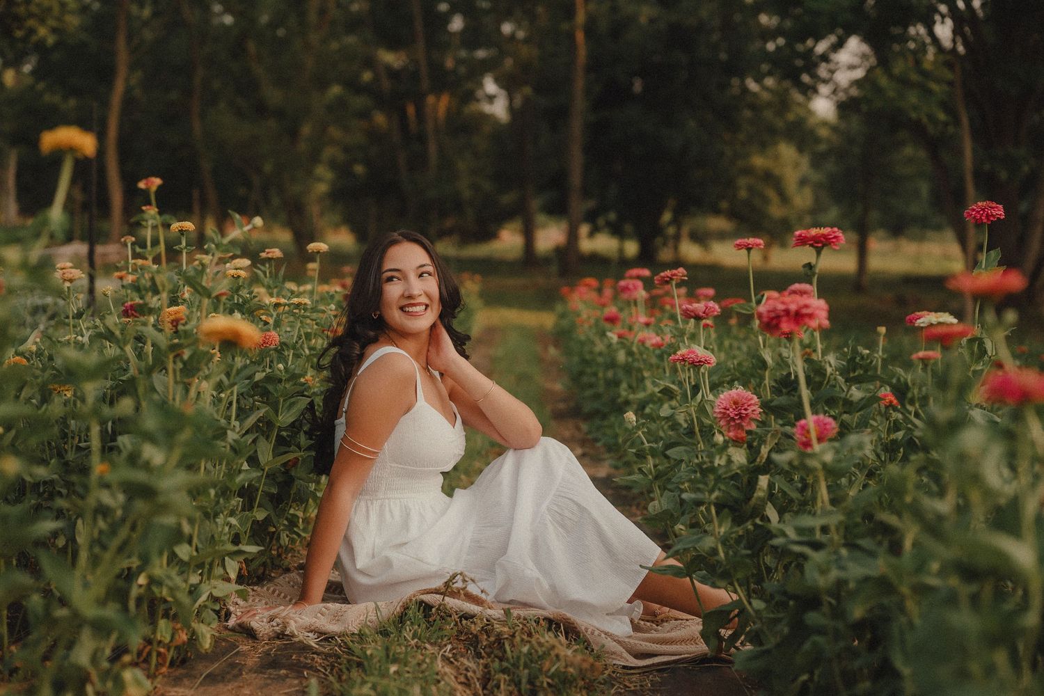 Woman in white dress sitting among colorful zinnia flowers in a garden, smiling warmly at golden hour.