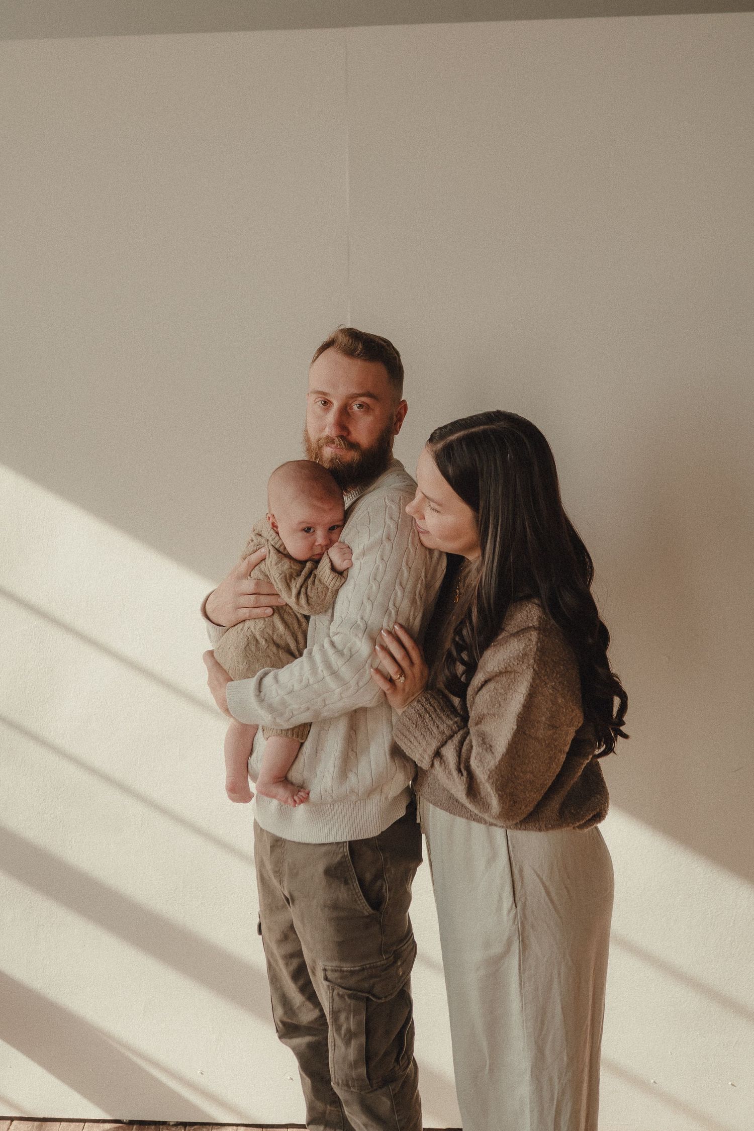 Minimalist family photoshoot against white wall with dramatic window lighting.