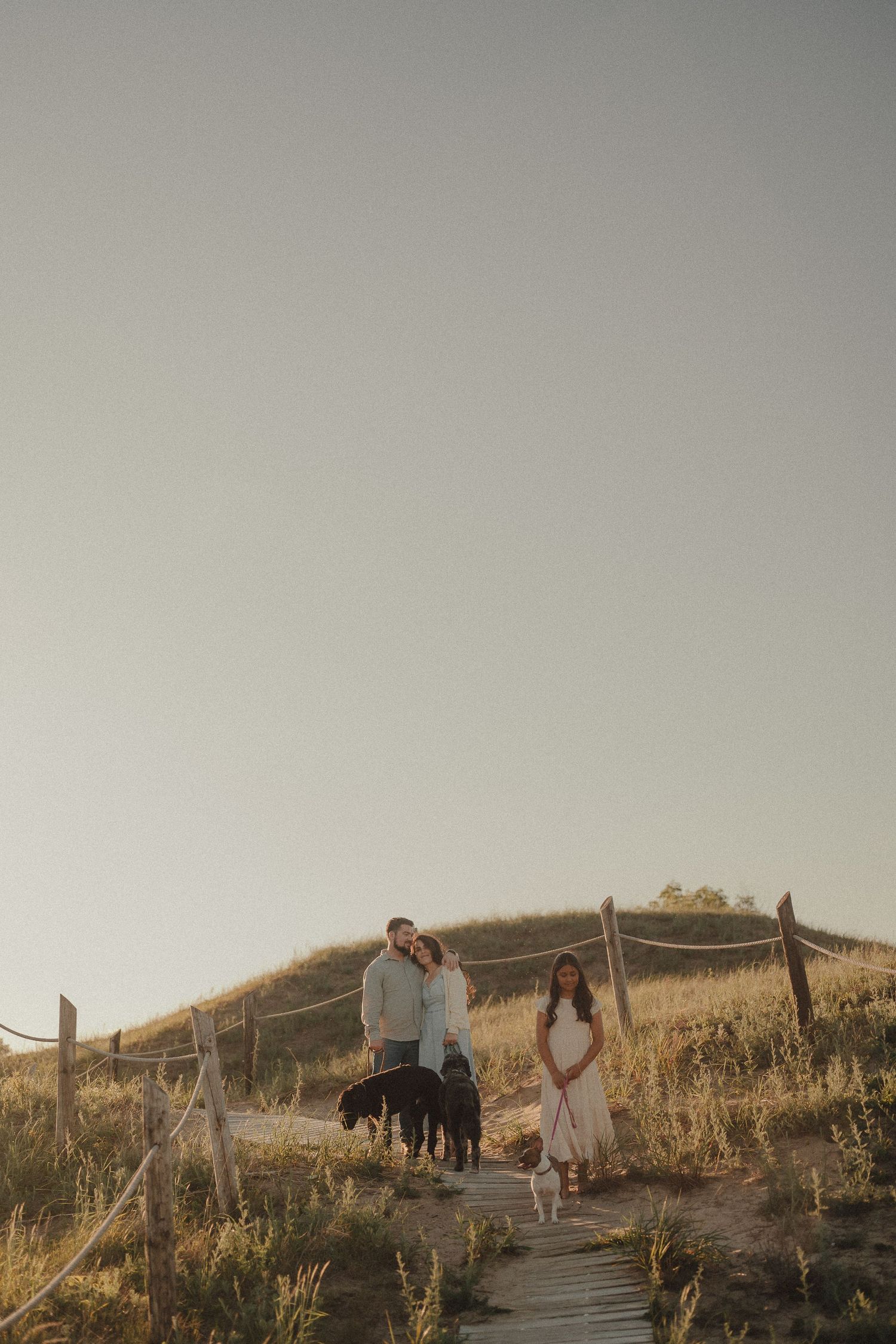 Couple with dogs on a wooden boardwalk path at golden hour in a scenic countryside setting.