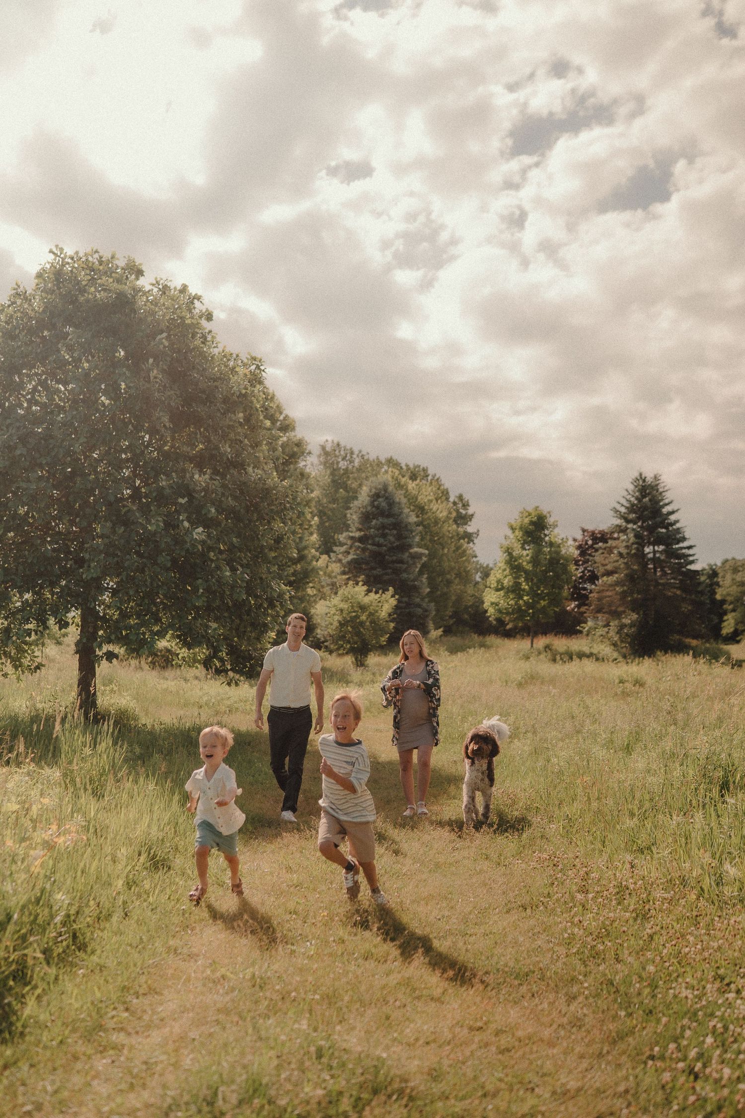 A joyful family with three young children runs through a sunny green meadow surrounded by trees.