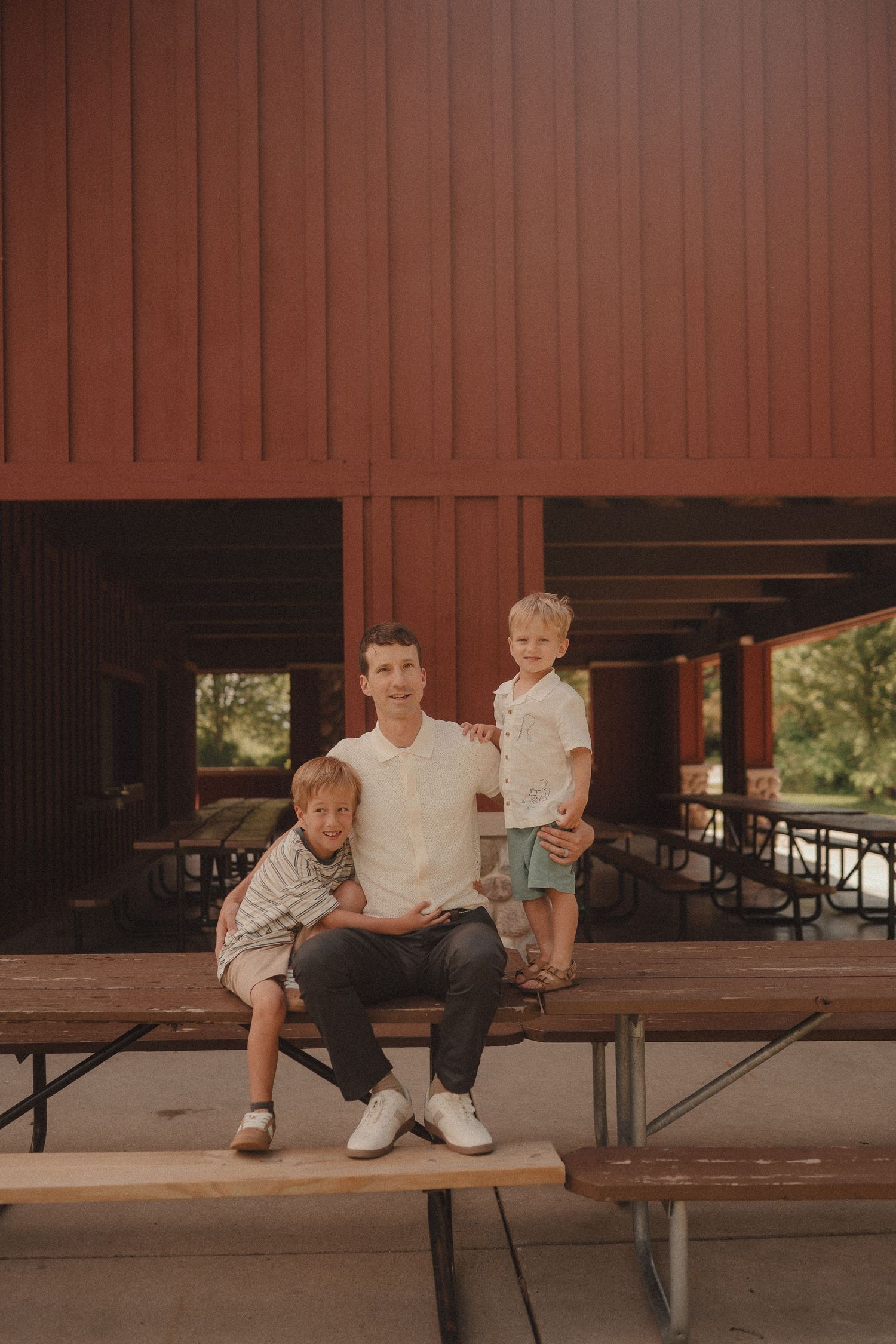 Father sitting on a picnic bench with two young sons in front of a red wooden building.