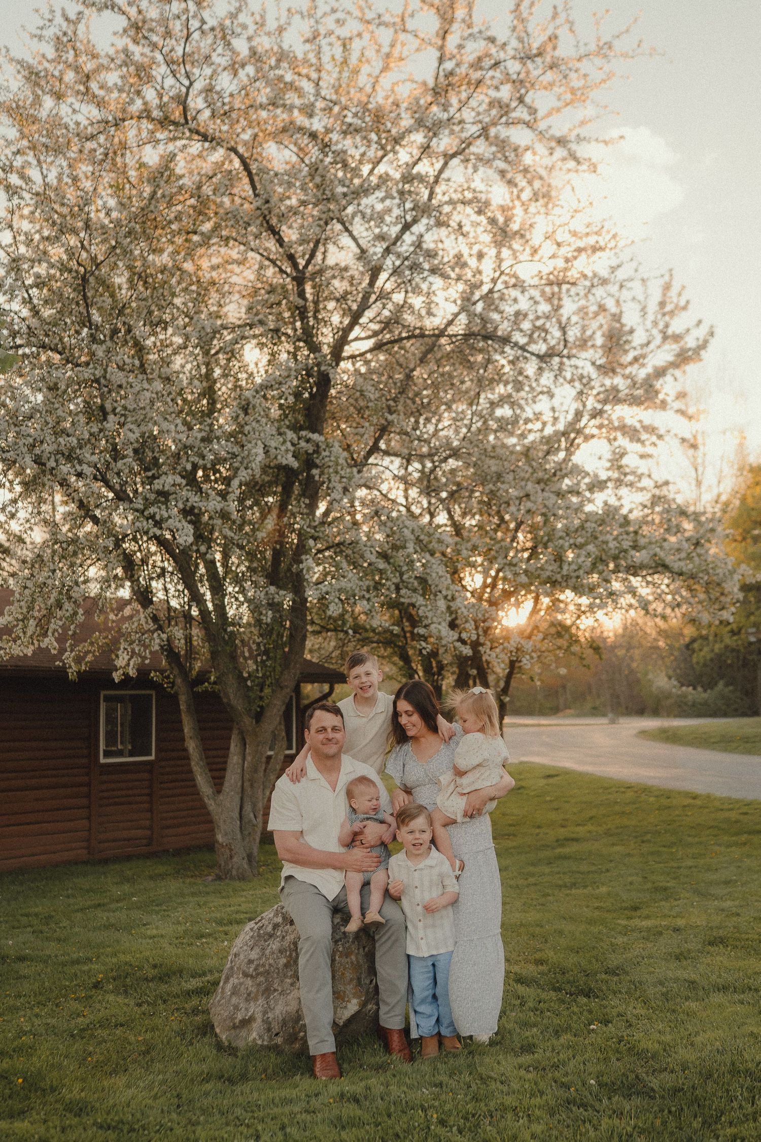 Family of five posing together at sunset in front of a blooming white tree near a rustic cabin.