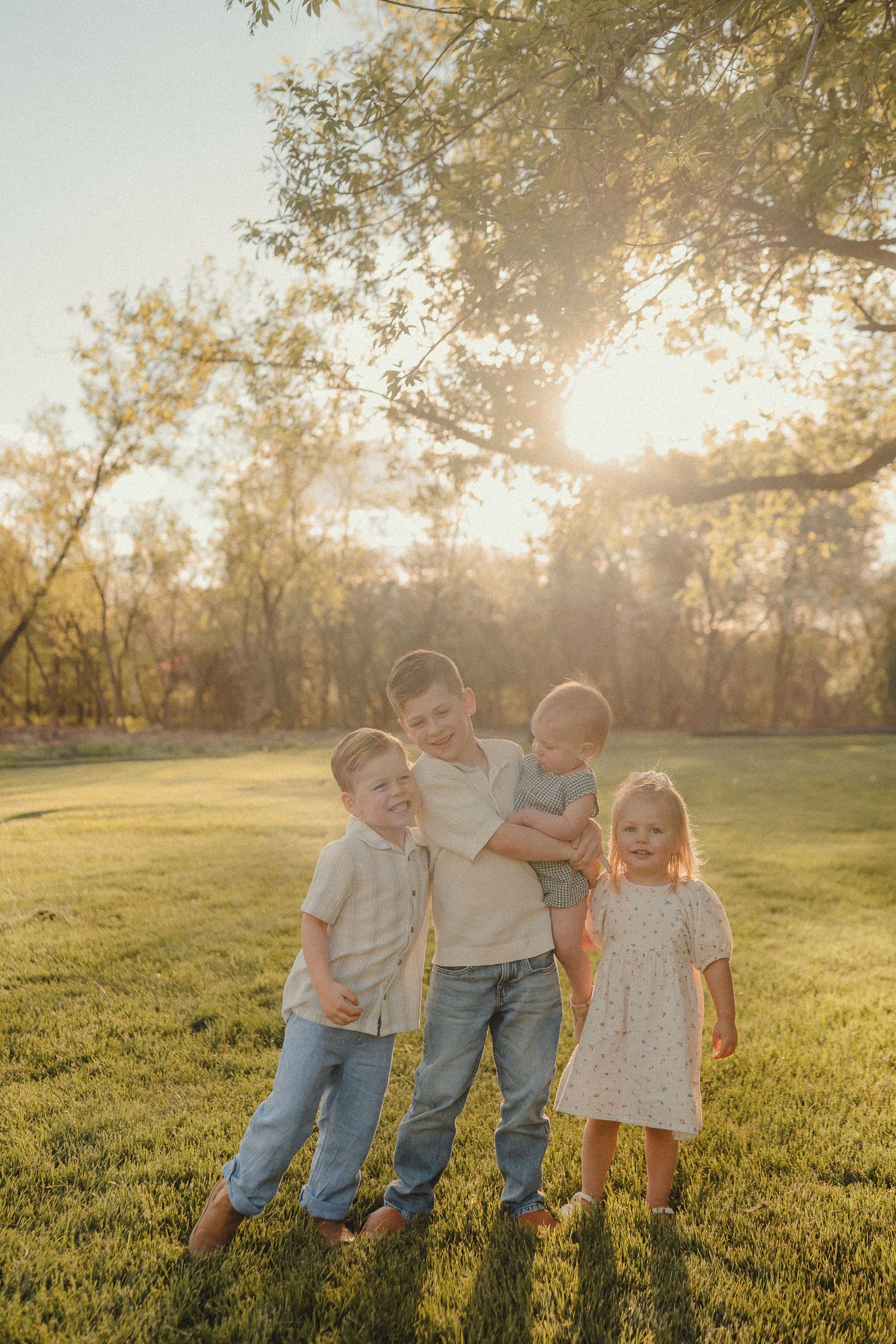 Four young siblings pose together on a sunny green lawn with golden backlight filtering through trees.