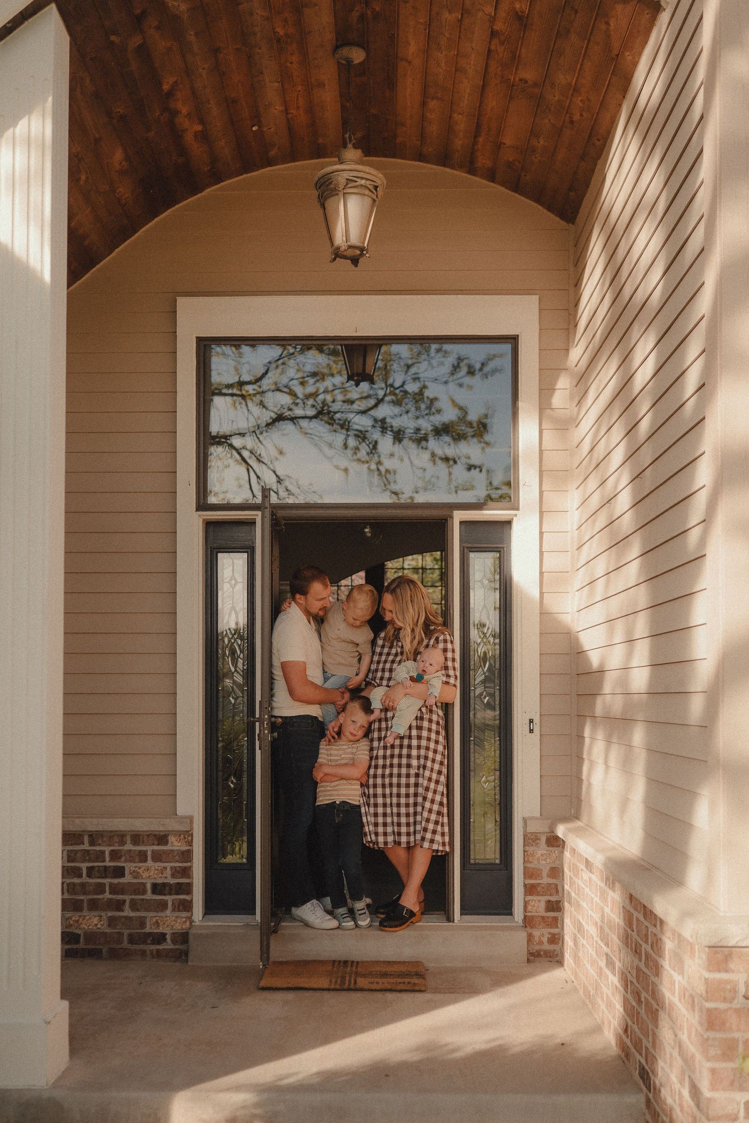 A family of four stands together smiling in the arched doorway of their brick home.