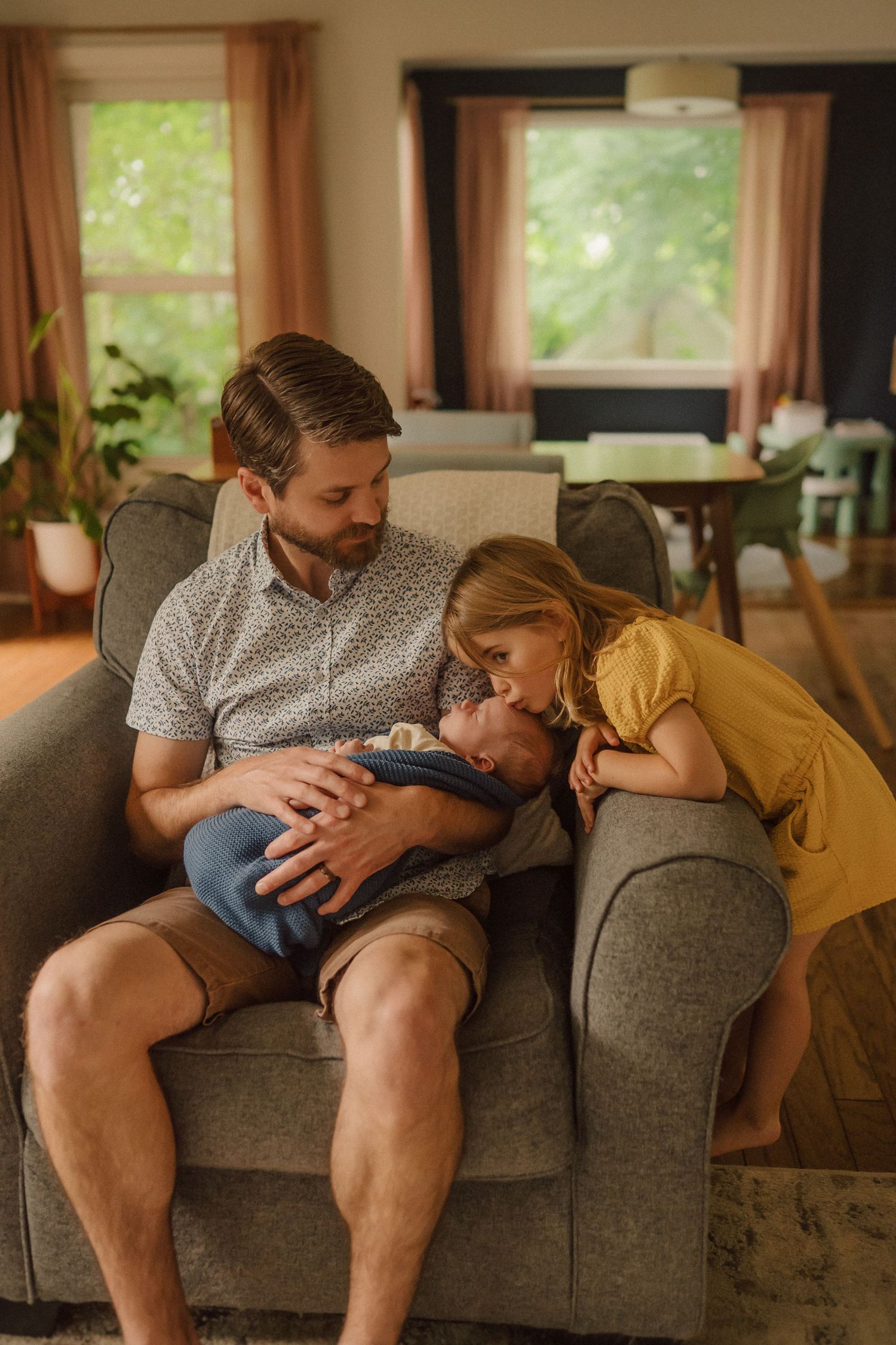 A tender family moment on a gray couch in a sunny living room with large windows.