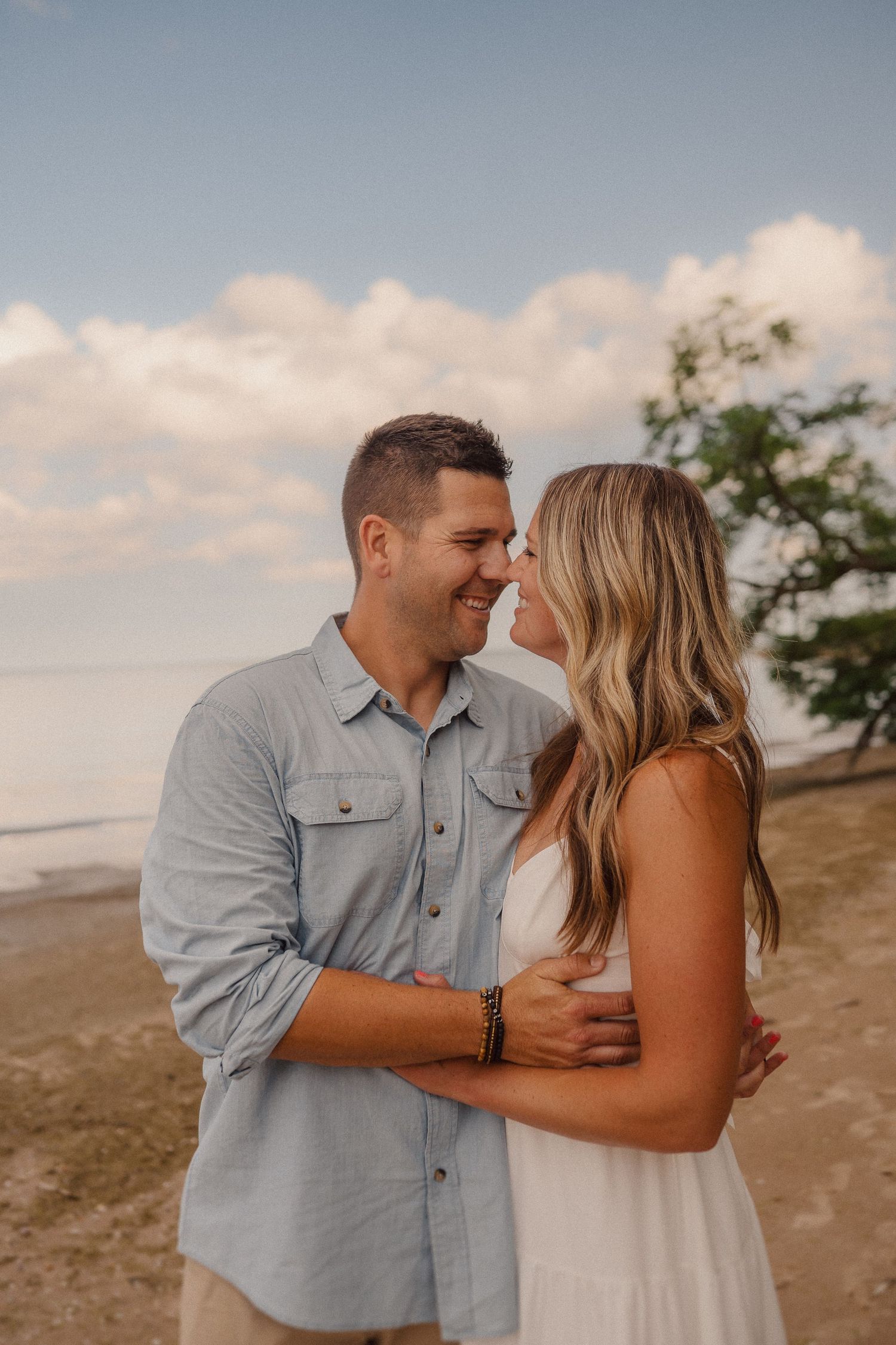 A couple shares an intimate moment on the beach with soft clouds and ocean backdrop during golden hour.