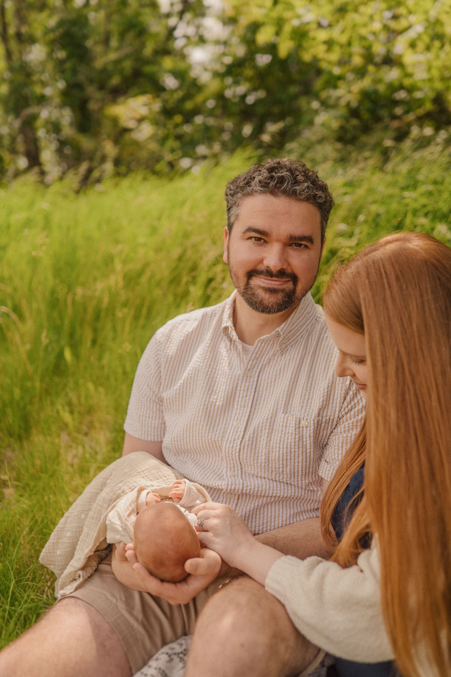 A couple sits together on grass outdoors with their newborn baby on a sunny summer day.