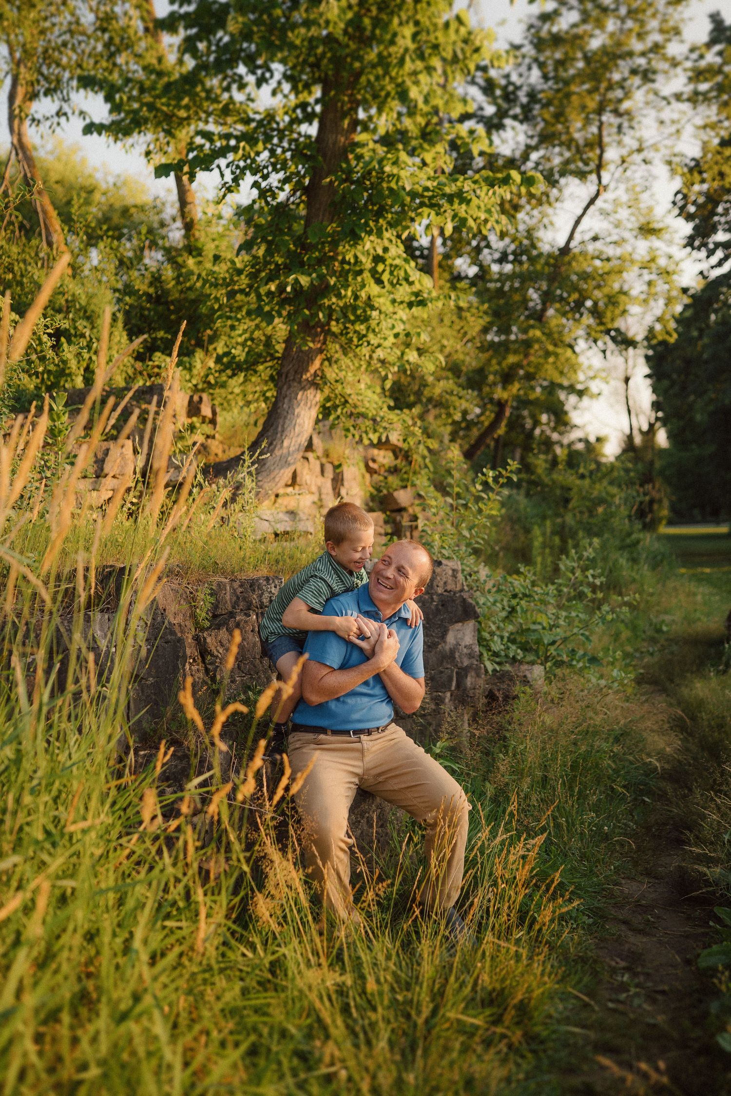 A tender moment outdoors in nature with sunlit trees and tall grasses creating a warm summer evening atmosphere.