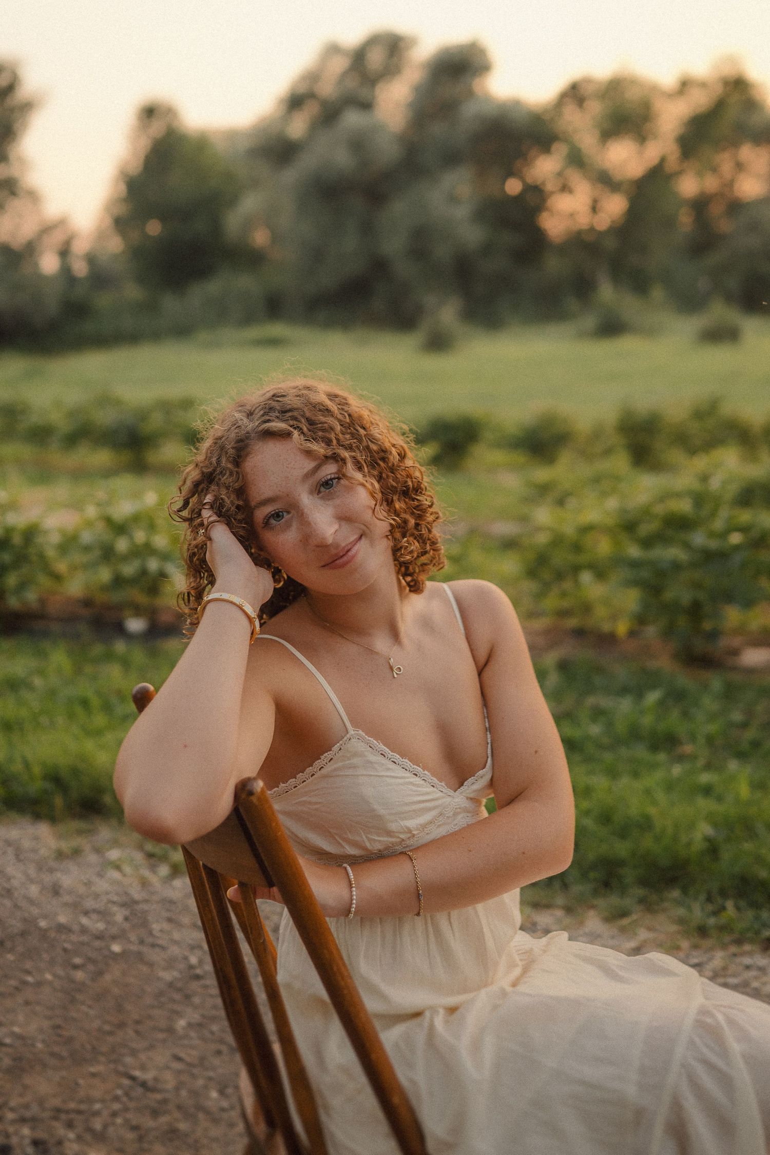 Ethereal outdoor portrait in a flowing white dress sitting on a wooden chair at golden hour in a grassy field.