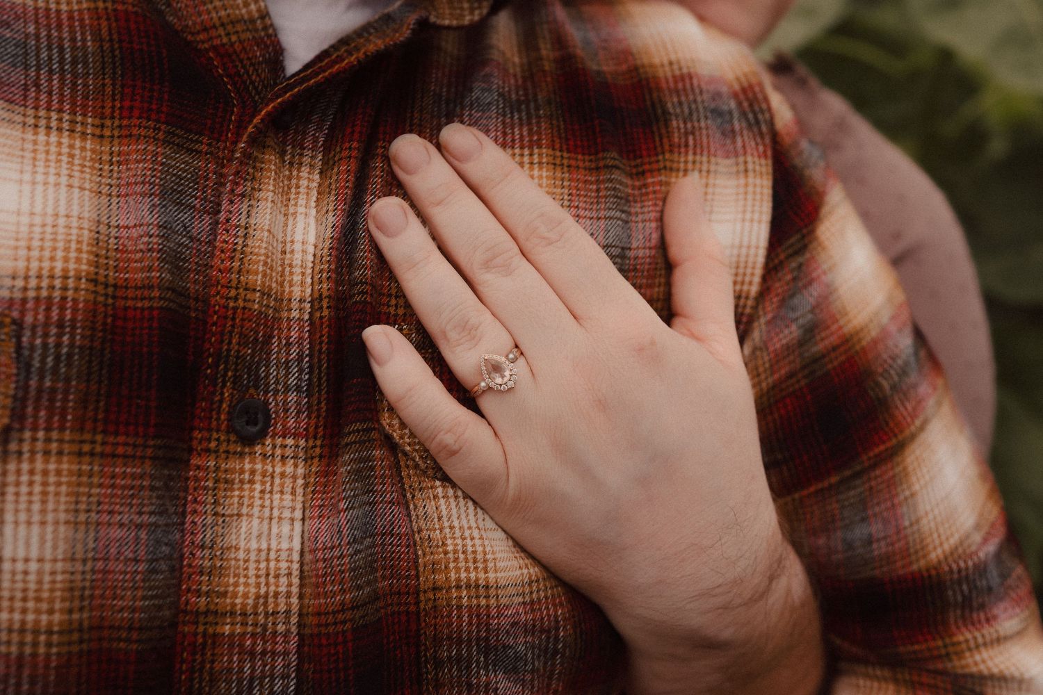 Close-up photo of an engagement ring on someone's hand against a plaid flannel shirt background.