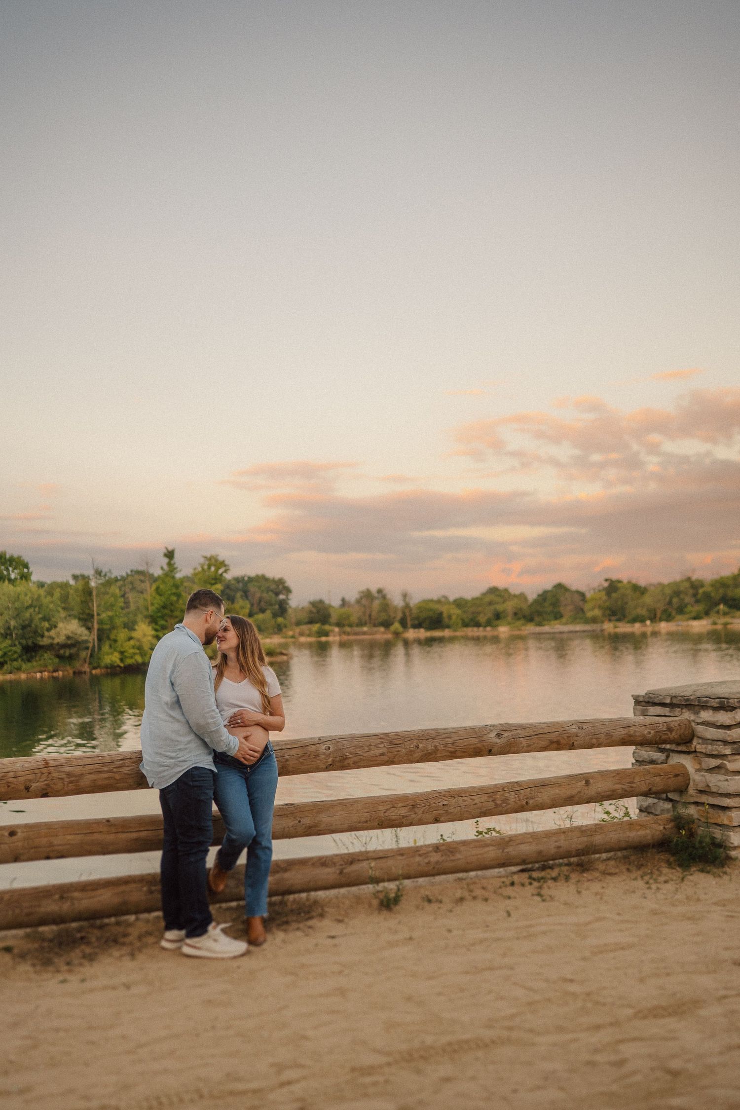 A couple shares an intimate moment at sunset while sitting on a wooden fence by a tranquil lake.