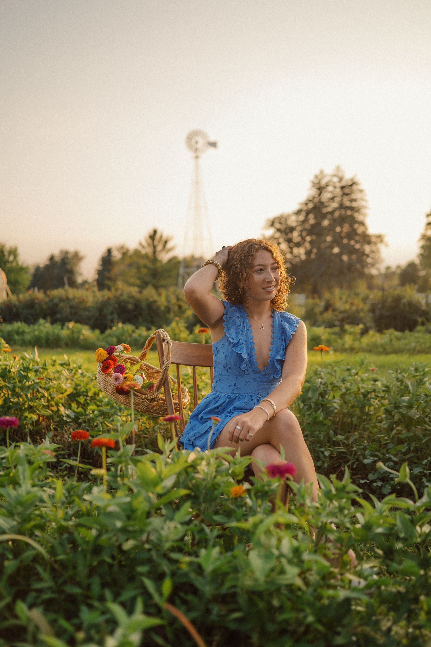 A woman in a blue dress sits gracefully in a vibrant garden filled with flowers at golden hour, with a windmill in the background.