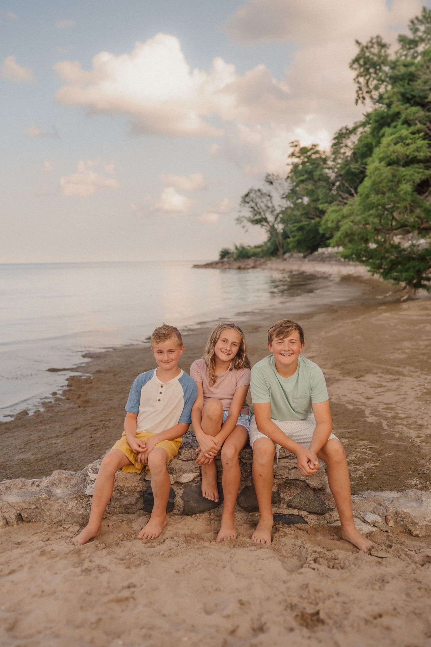 Three children sit together on rocks at the beach during sunset with trees and ocean in the background.