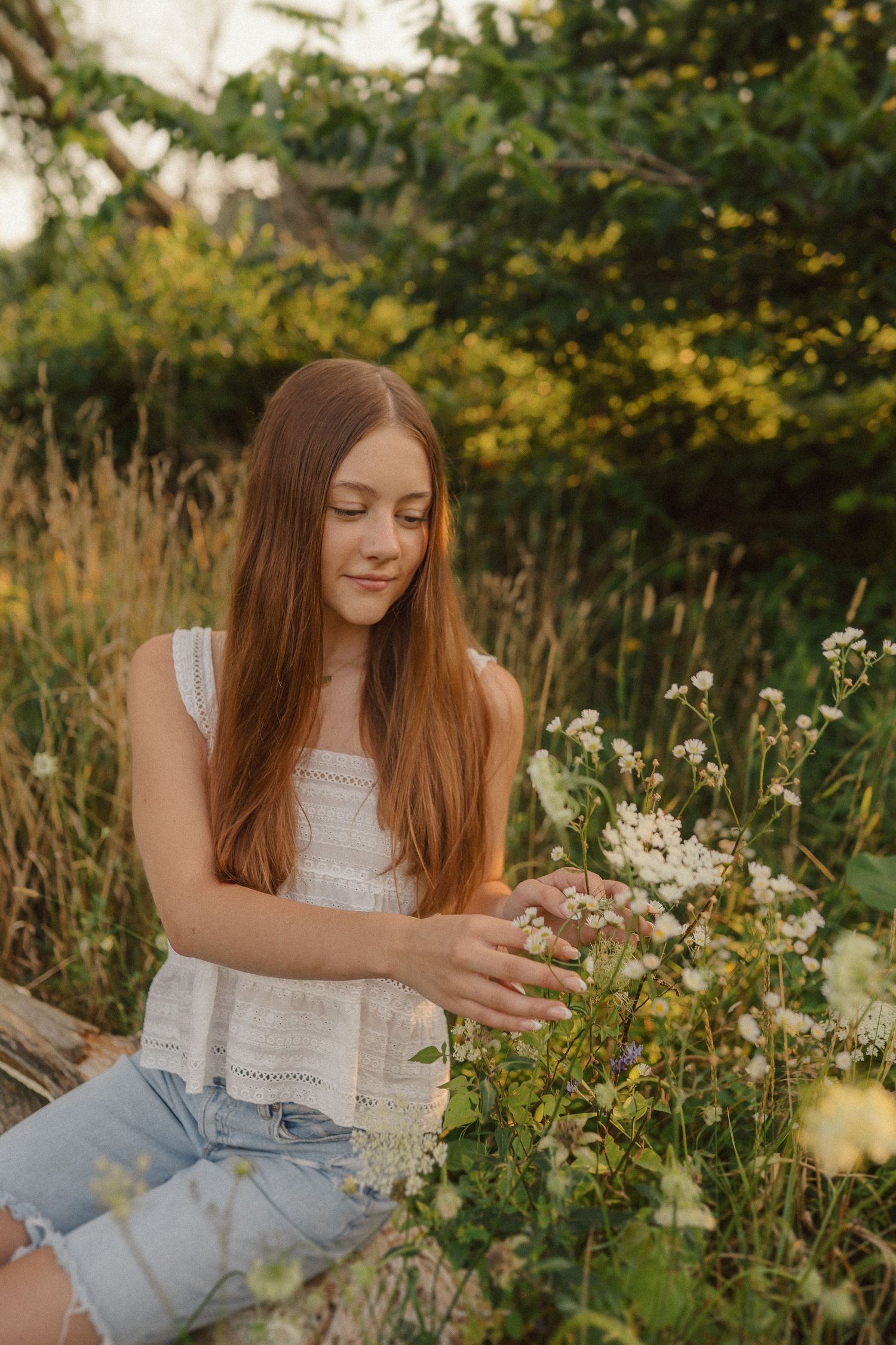 A person in casual attire reaches out to touch delicate white wildflowers in a garden setting at golden hour.
