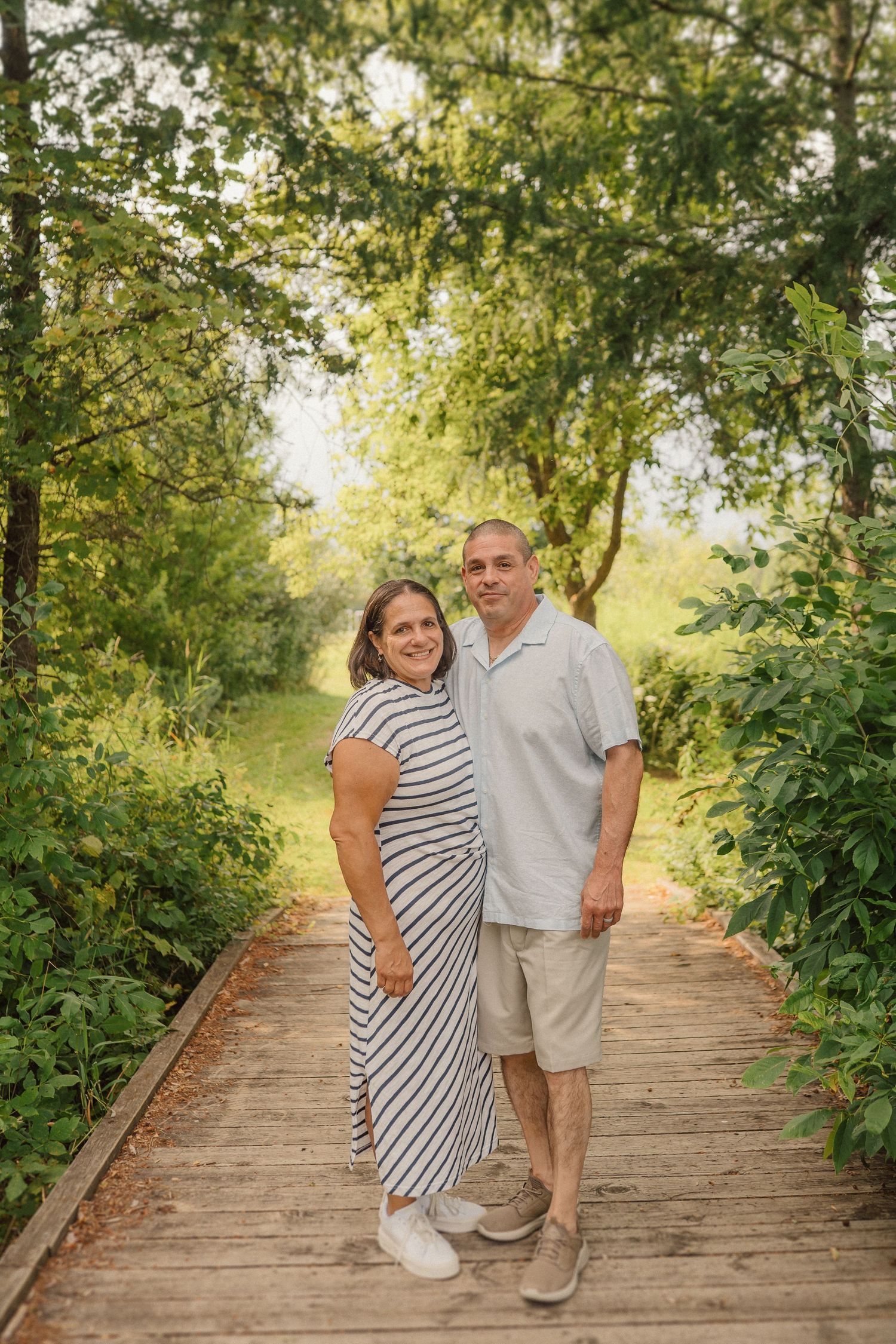 Couple poses together on a wooden bridge surrounded by lush green trees and natural lighting.