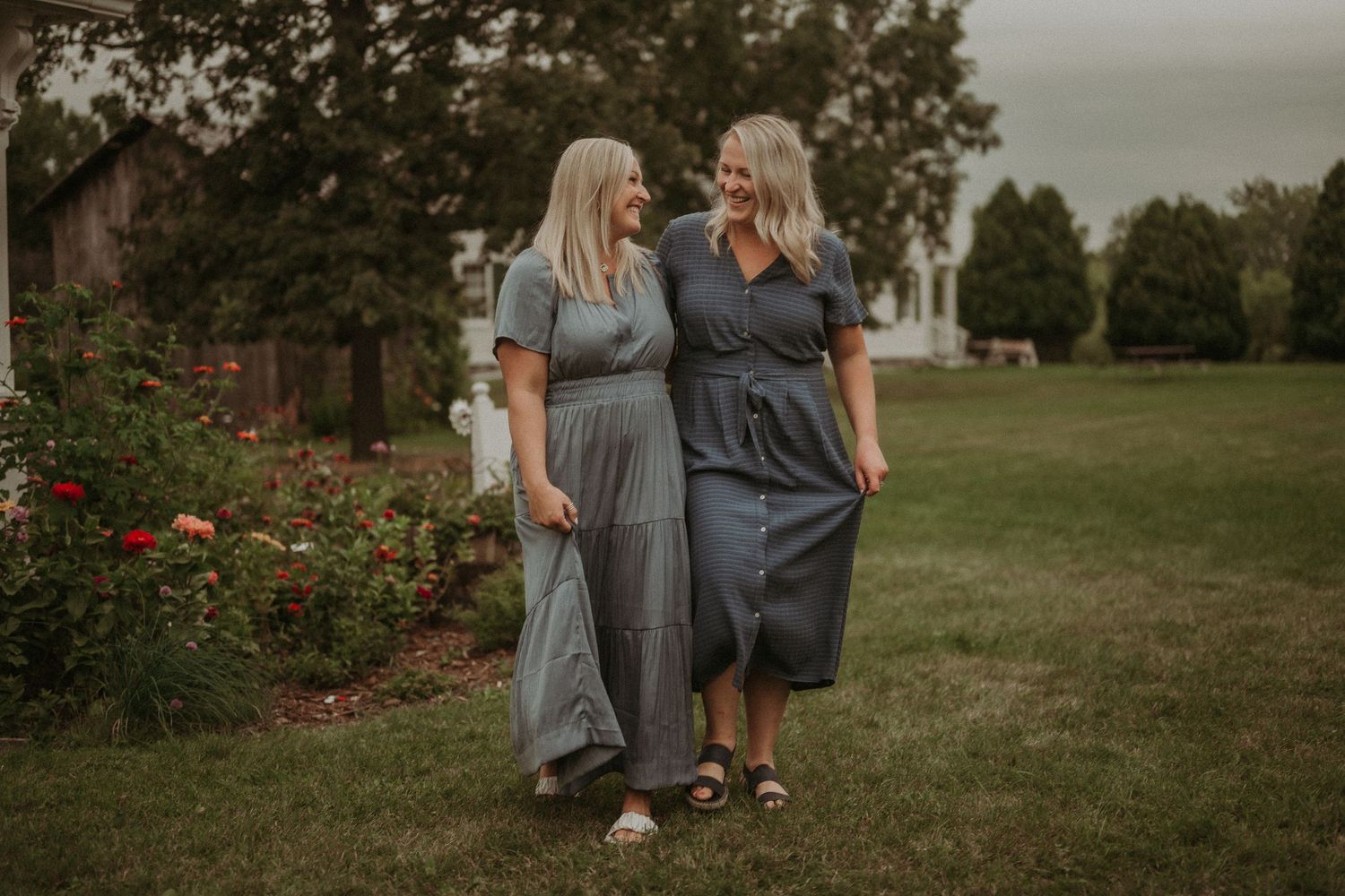 Two people in grey dresses walking together through a garden path with blooming flowers.