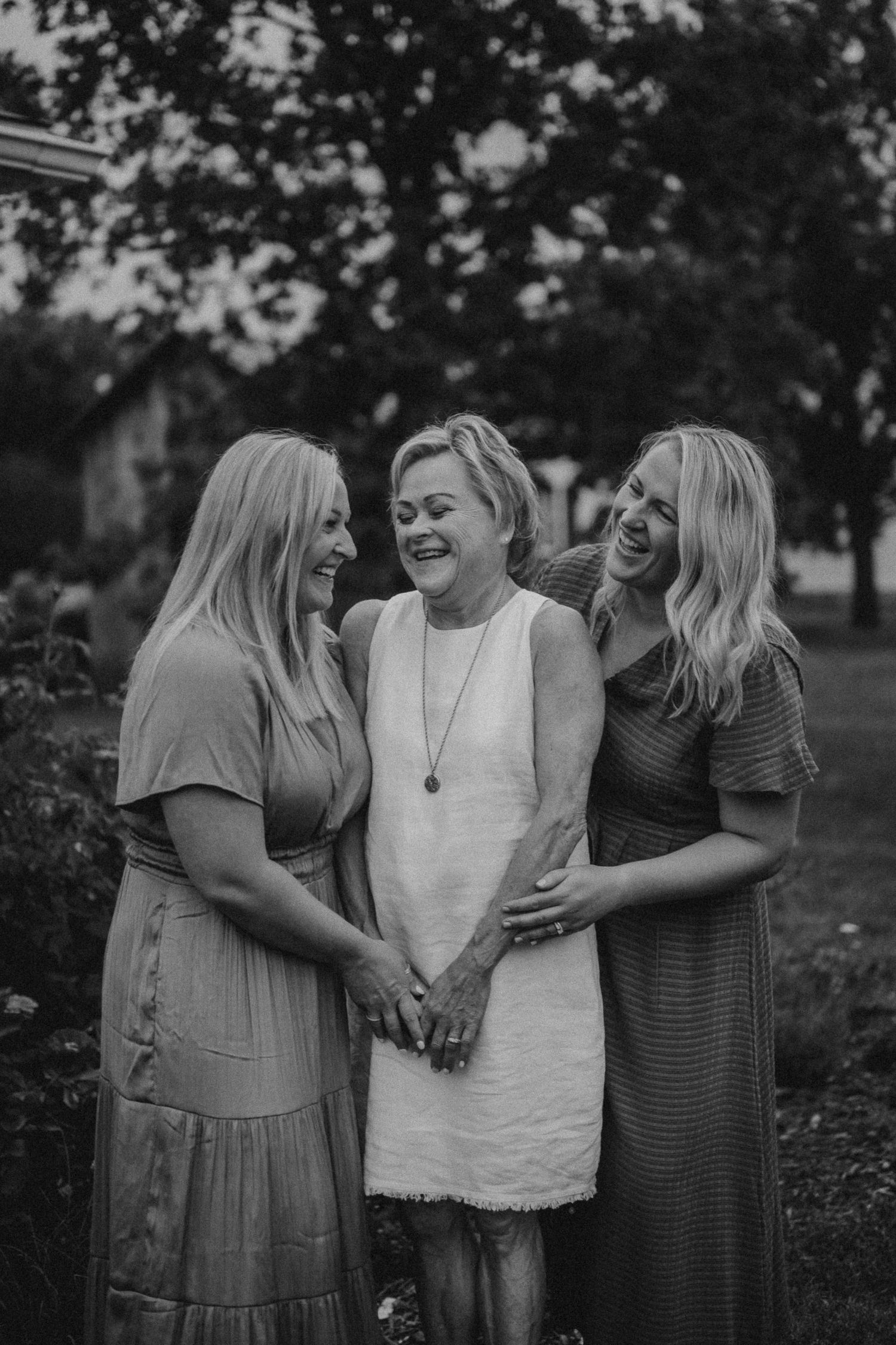 Black and white portrait of three women embracing and laughing in garden setting.