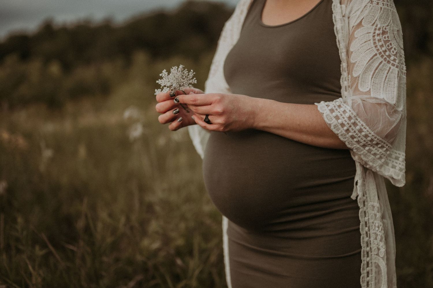 Pregnant person in a brown dress holds a small flower while standing in a field at sunset.