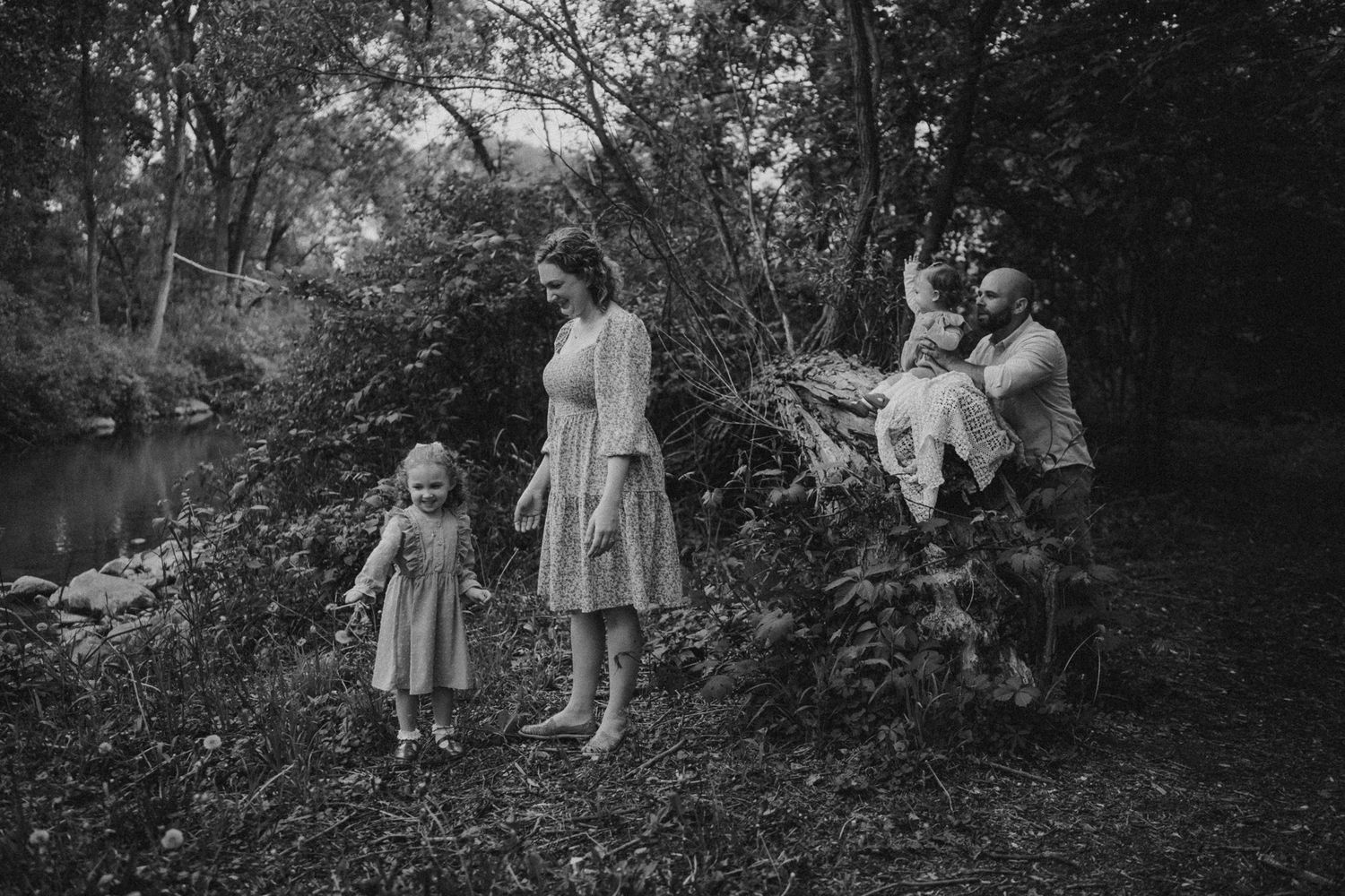 A black and white vintage style photo of a family spending time together in a wooded area near a stream.