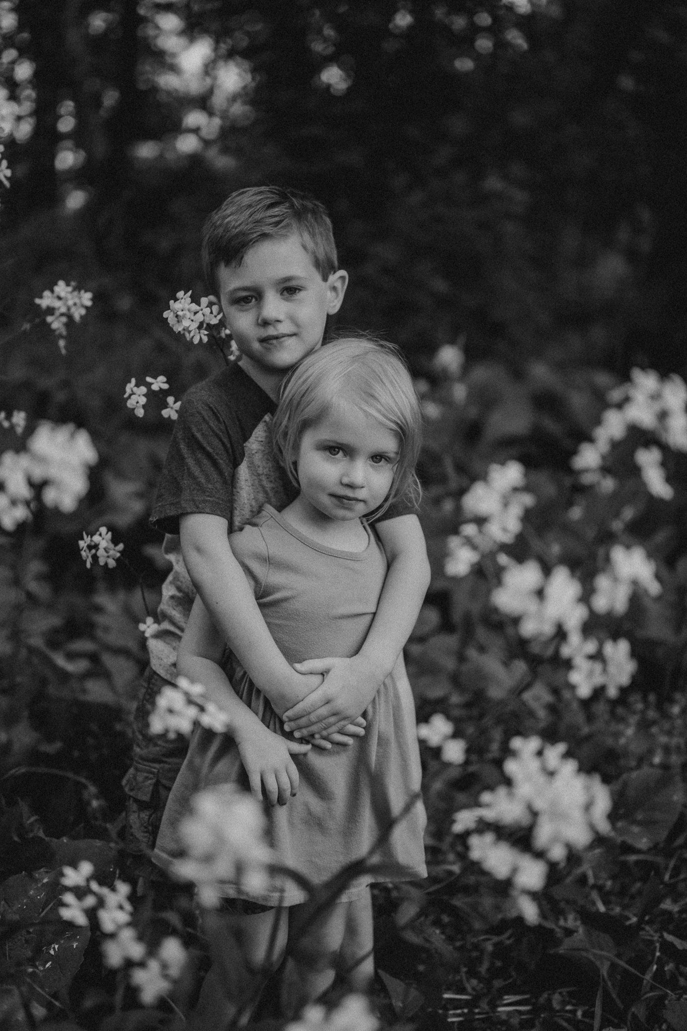 Black and white portrait of two children hugging amongst blooming flowers outdoors.