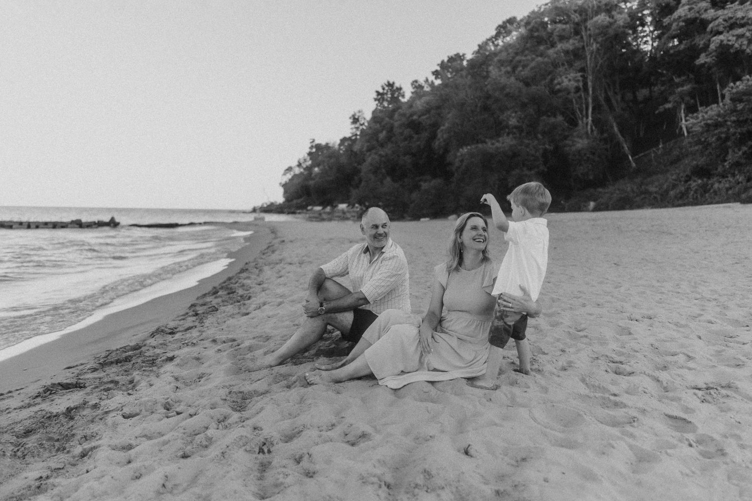 A peaceful beach scene with three figures sitting on the sand near wooded shoreline in black and white.