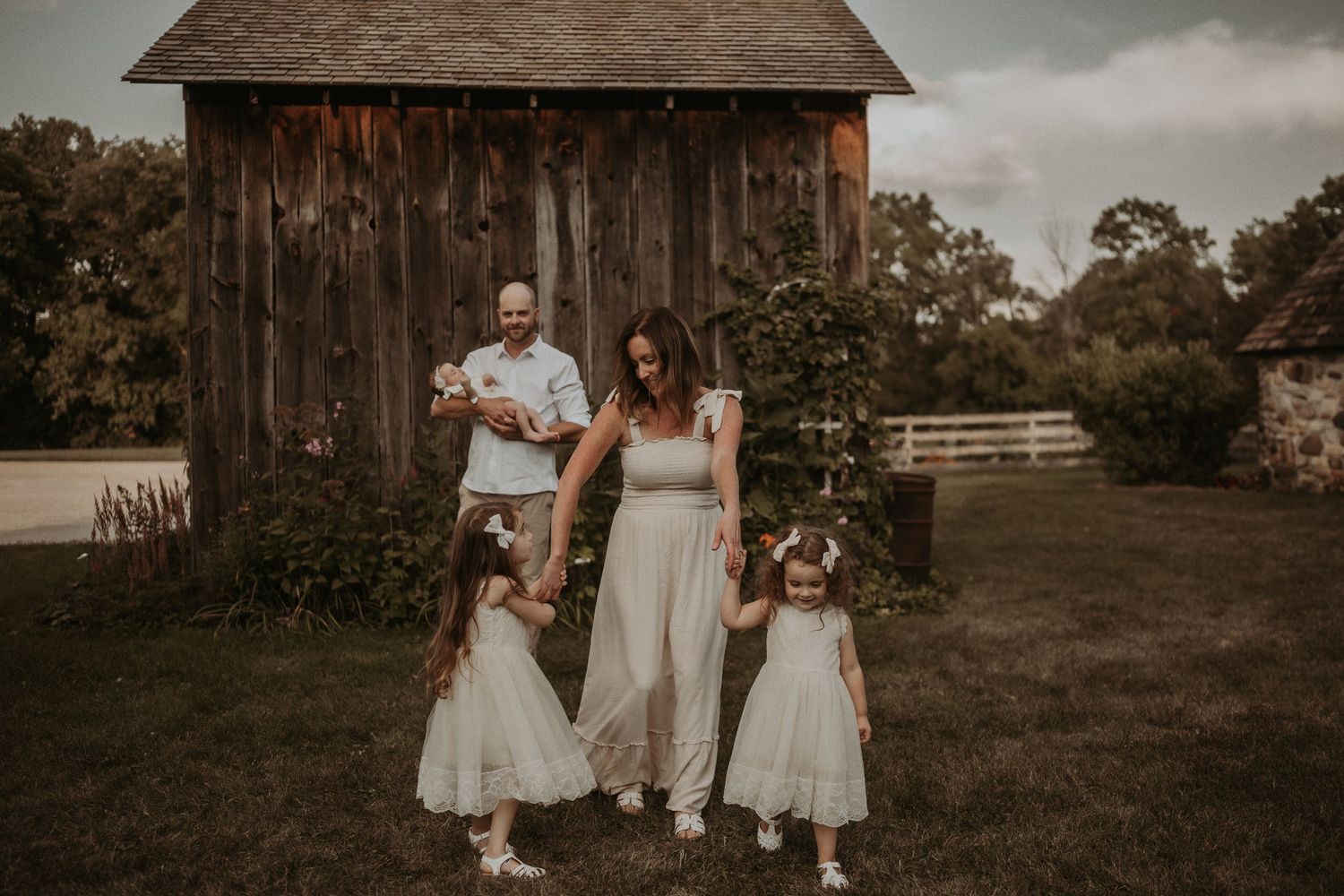 Rustic family portrait in front of a weathered barn with matching white outfits in a countryside setting.