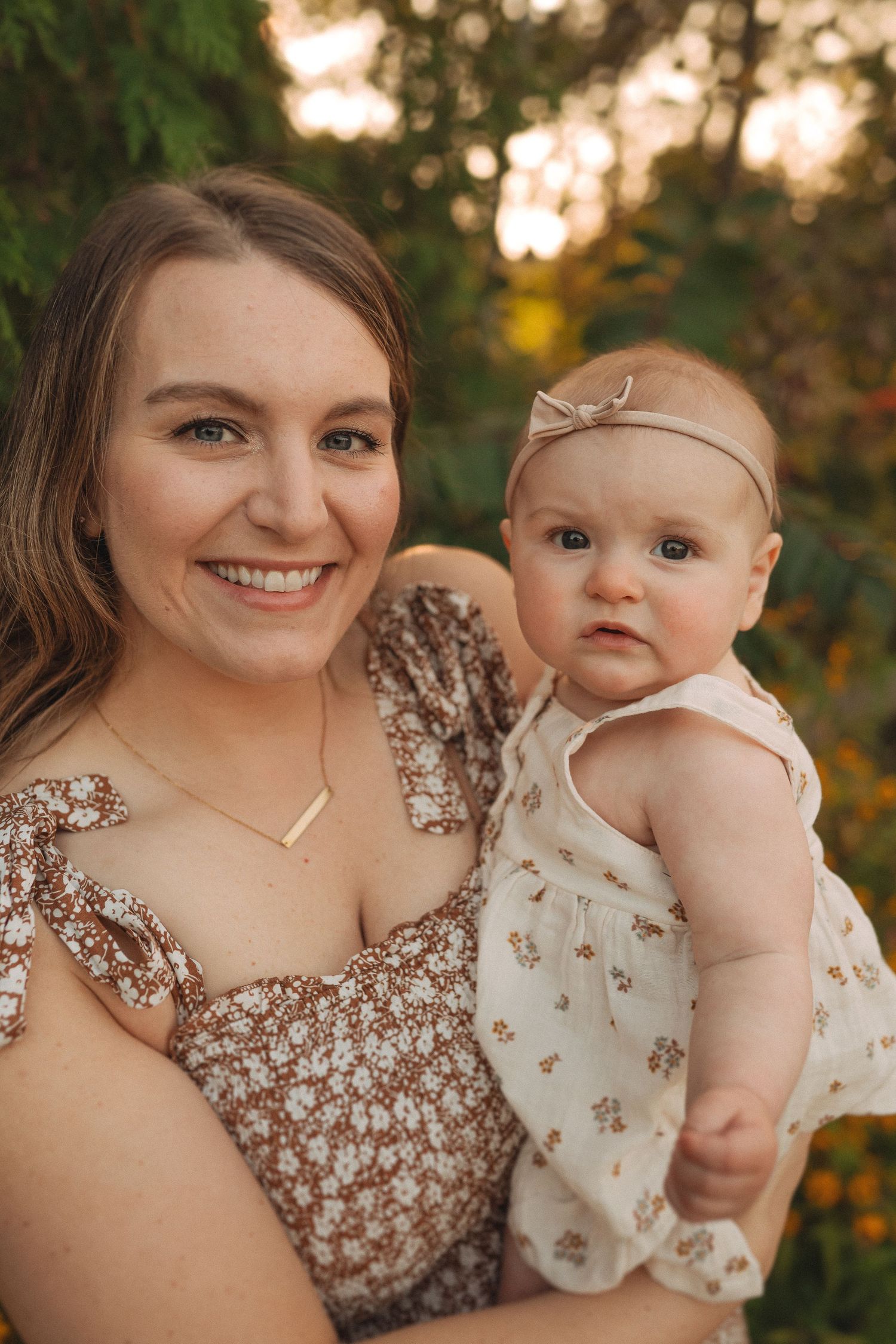 Close up portrait in a floral dress with infant in white dress against autumn foliage.