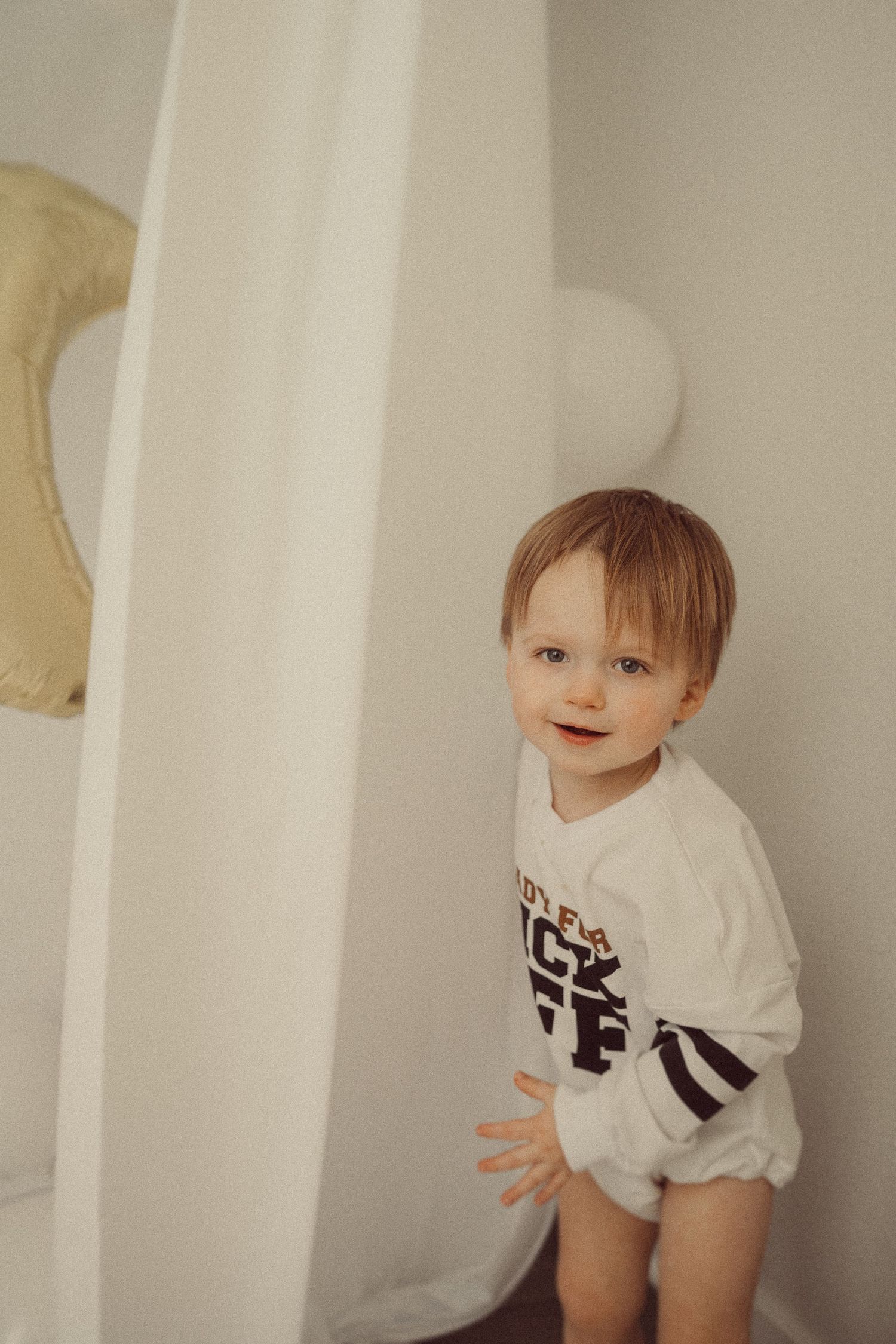 Toddler in white sports onesie standing against white wall with gold moon decoration.