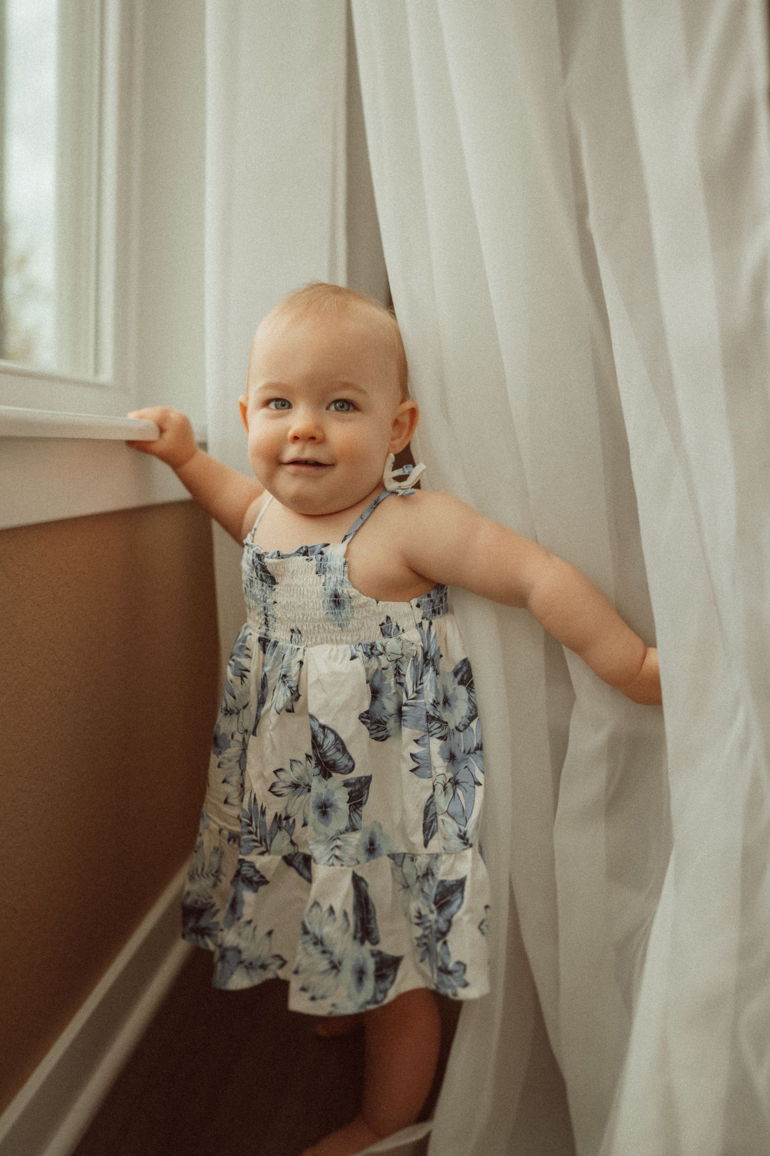 Baby stands by window wearing blue floral dress while playing with white curtains.