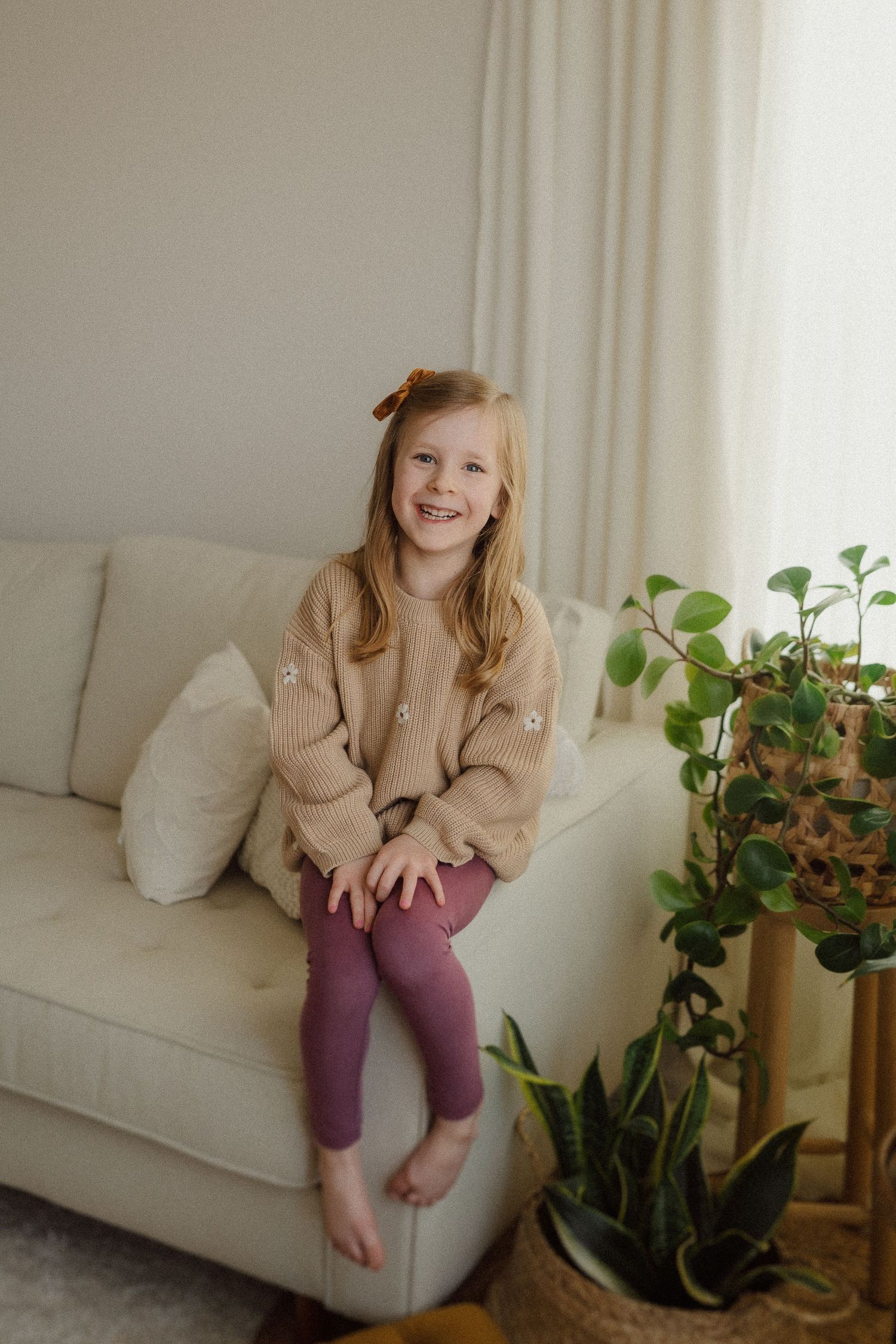 Child wearing tan sweater sits on white couch beside houseplants with natural lighting from window.