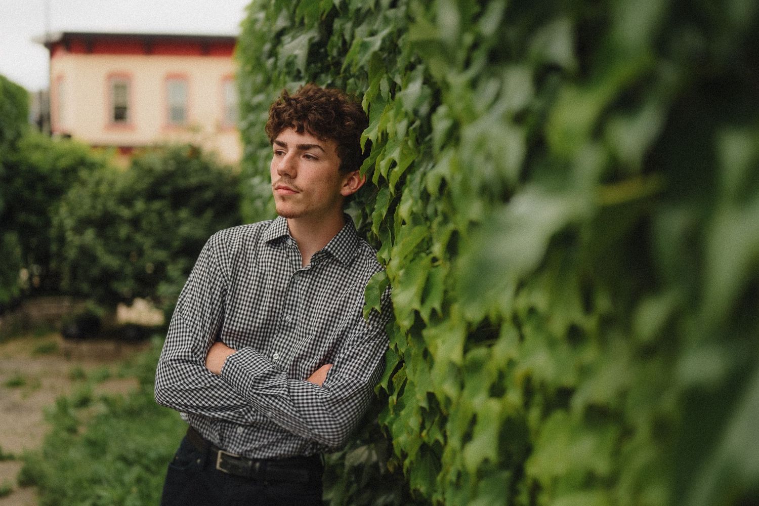 A person in a checkered shirt stands with arms crossed near lush green vines on a sunny day.