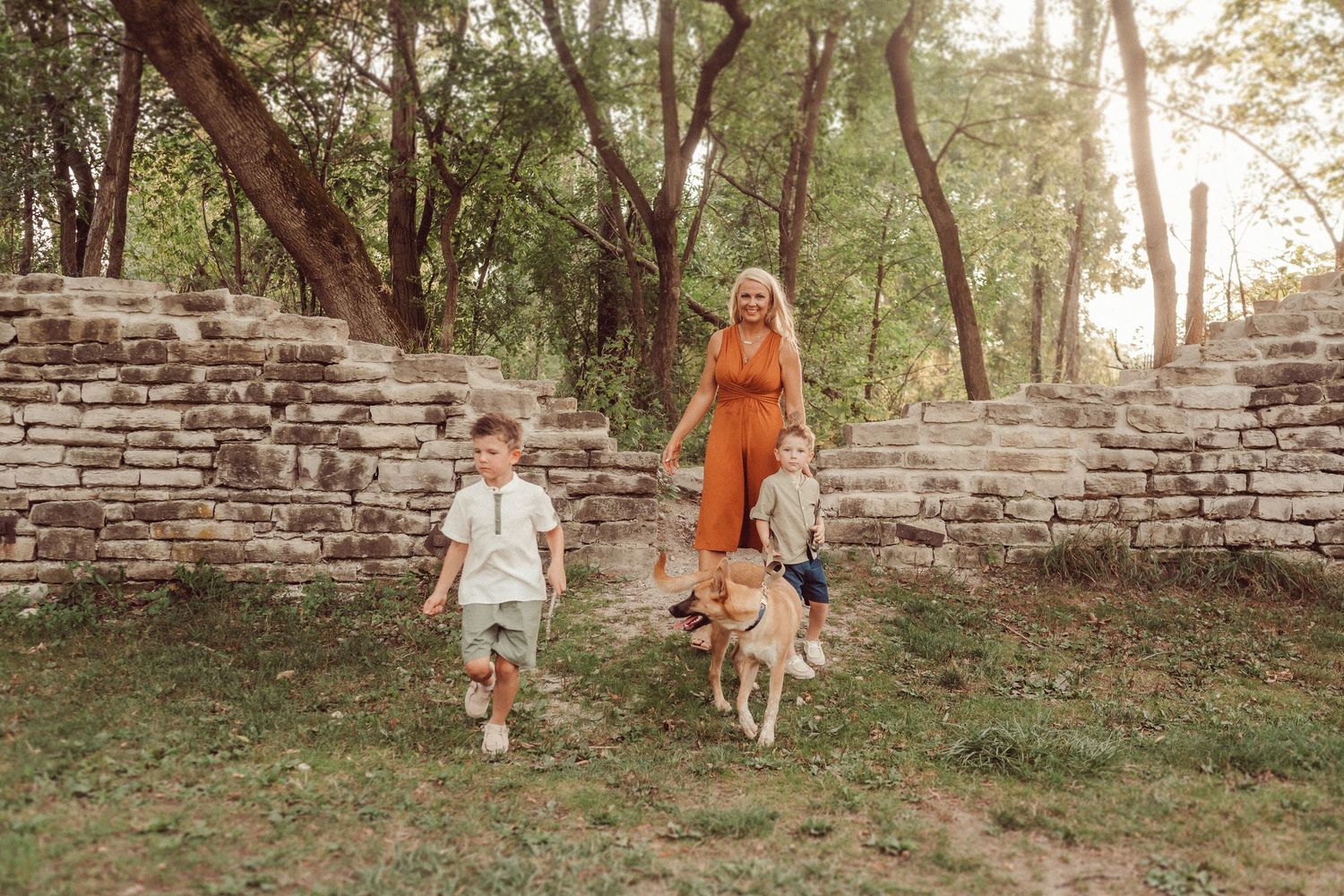 A family walks along stone walls with their dogs in a scenic wooded area.