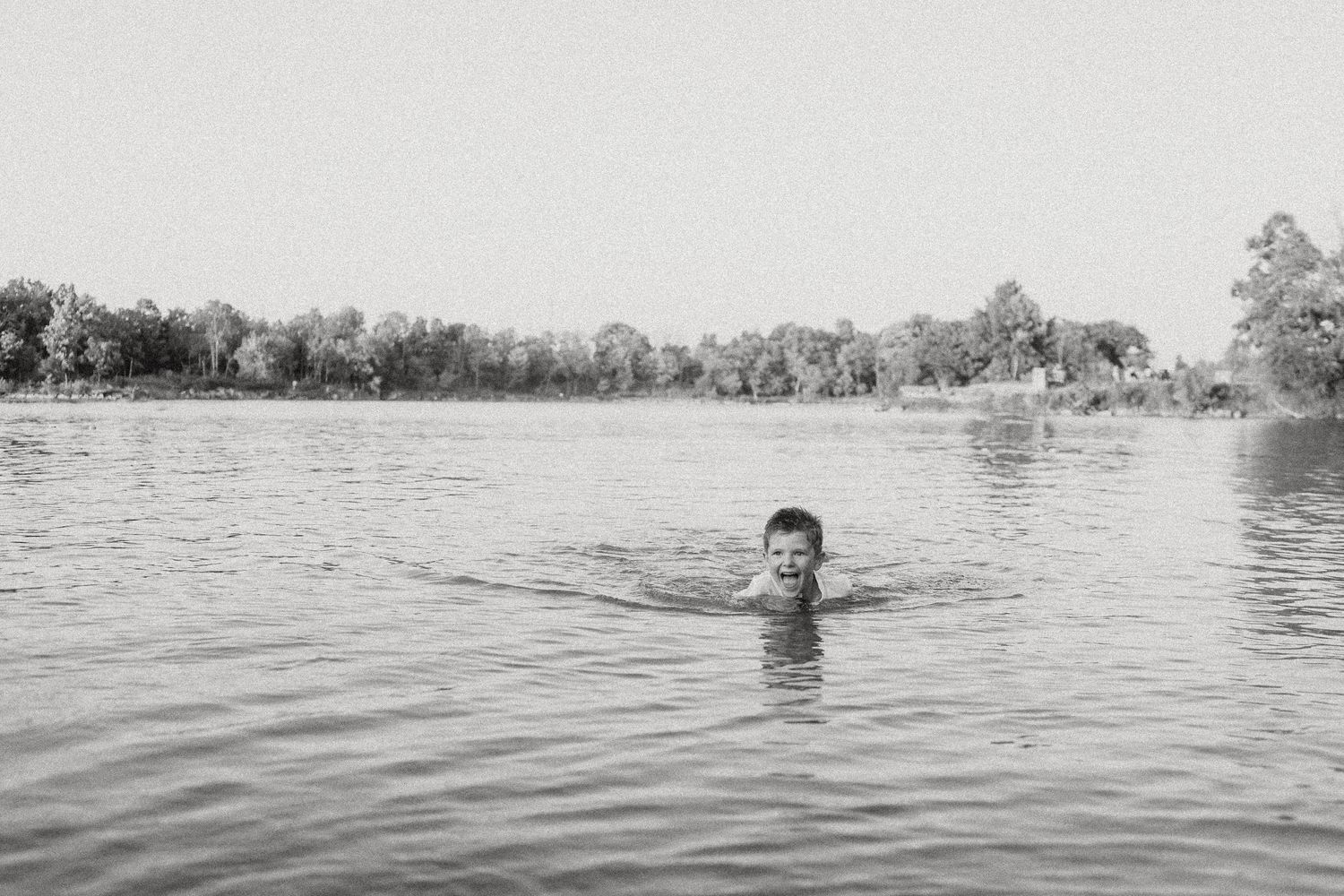 Black and white photo of someone swimming in a peaceful lake surrounded by trees.
