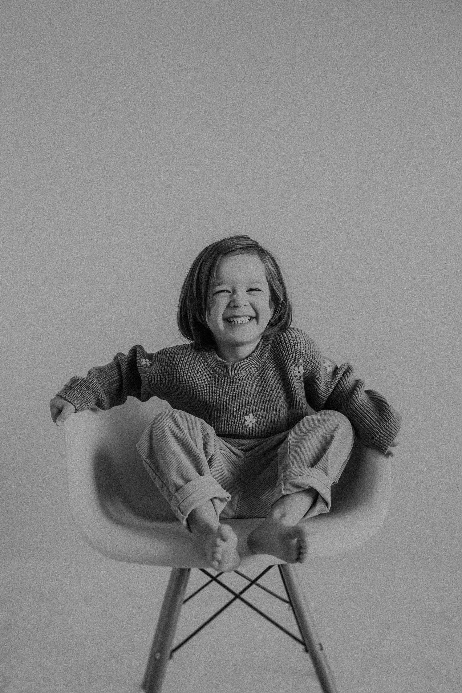 Child with bright smile sits casually in white modern chair in monochrome portrait.
